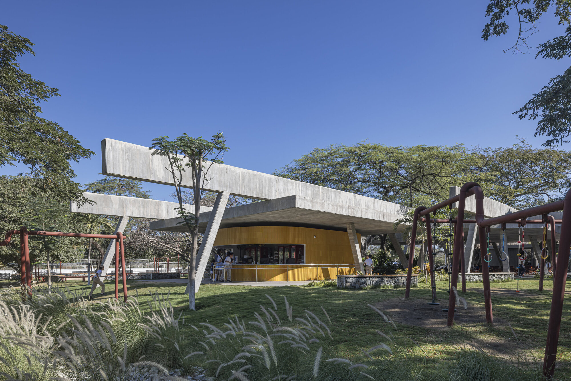 Yellow curved kiosk wall beneath a concrete roof on slanted supports, seen across a lawn with grasses and a red swing set