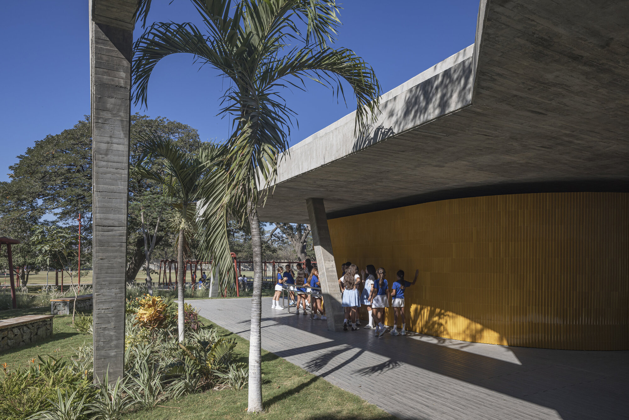 Group of schoolgirls standing beside the curved yellow tile wall under the concrete roof edge, with palm trees in bright sun