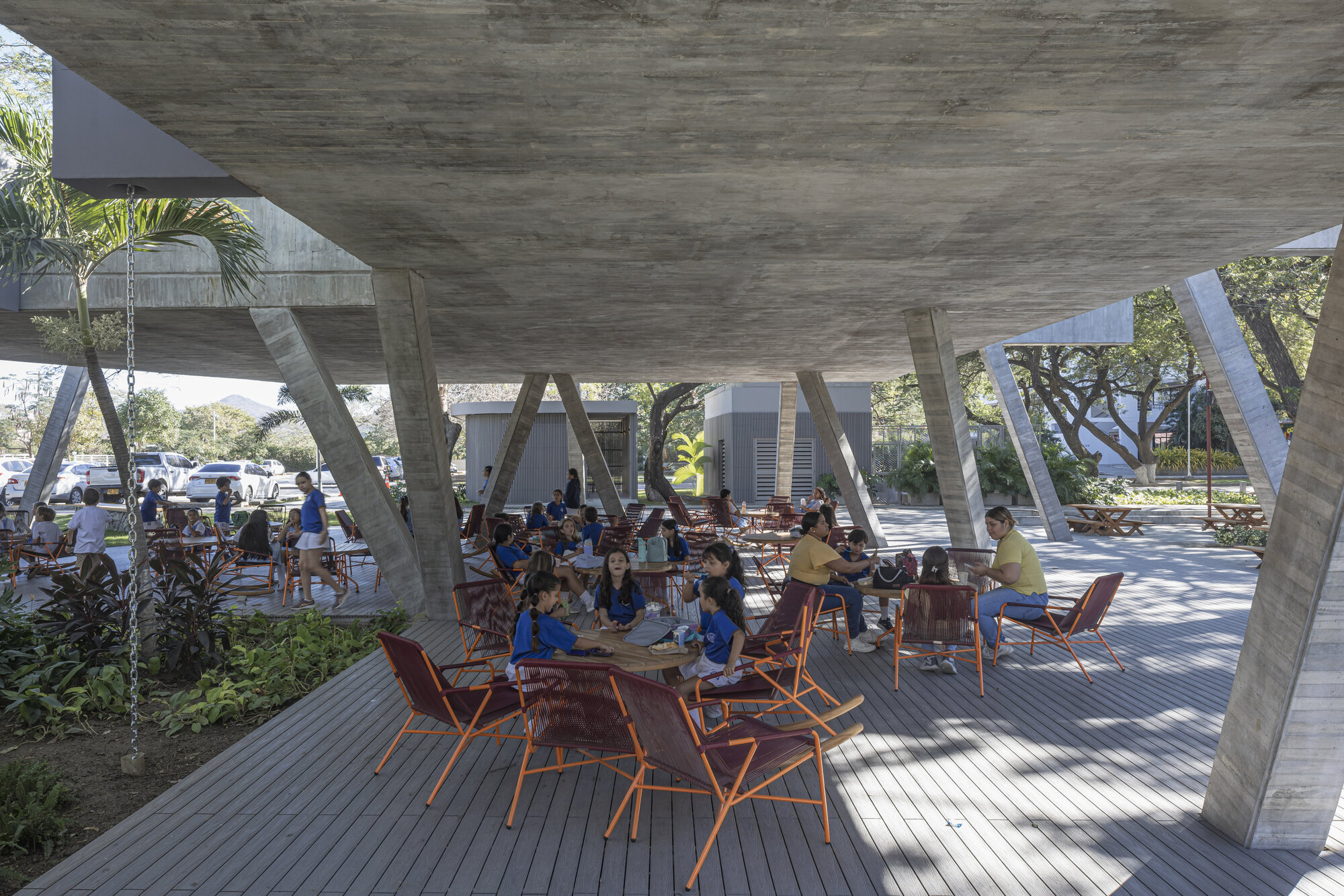 Underside of the board-marked concrete slab with tilted columns above a timber deck where children and adults sit at tables