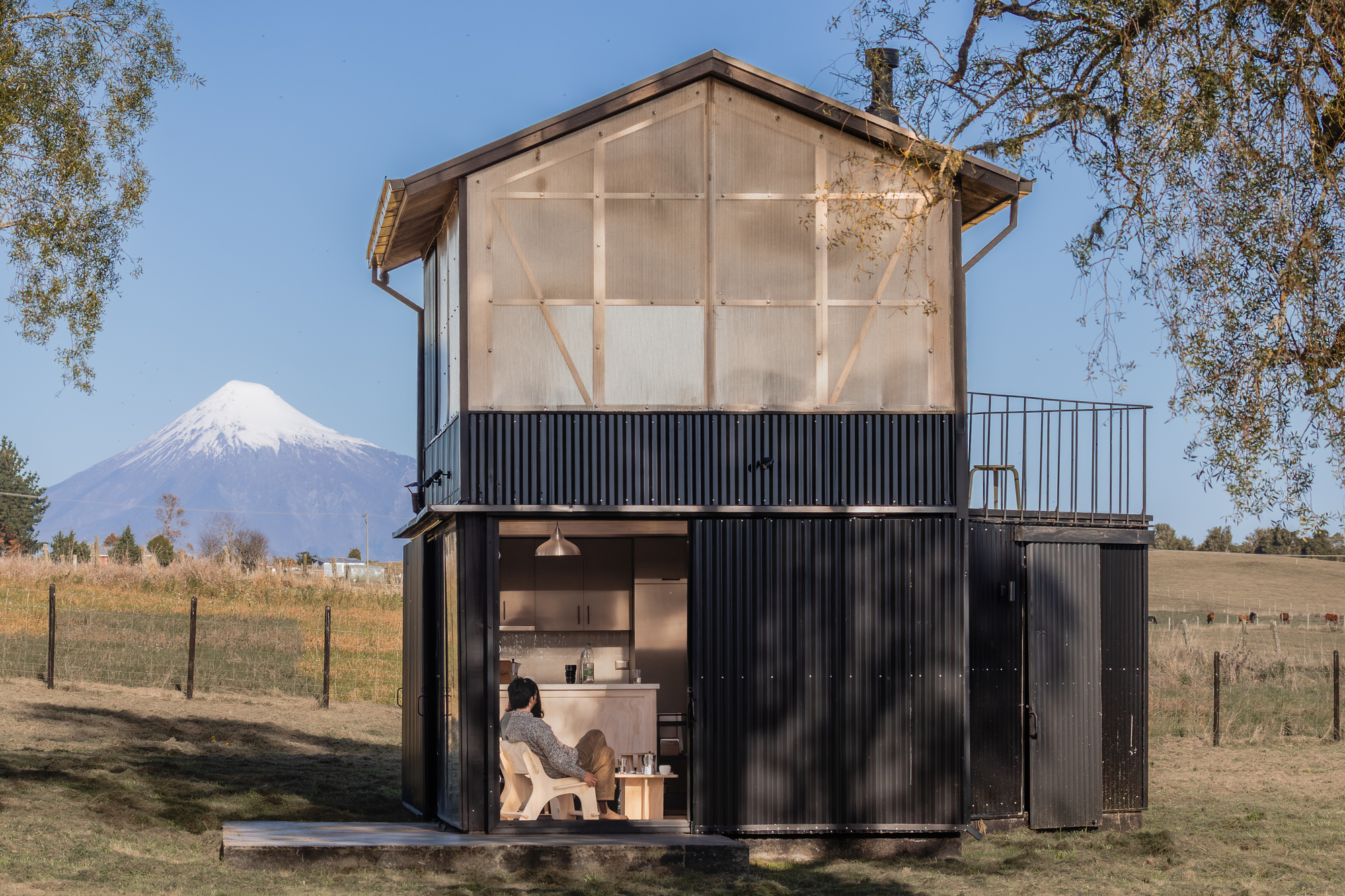Two-storey black corrugated cabin with translucent upper wall and open kitchen, a snow-capped volcano rising behind in clear afternoon light
