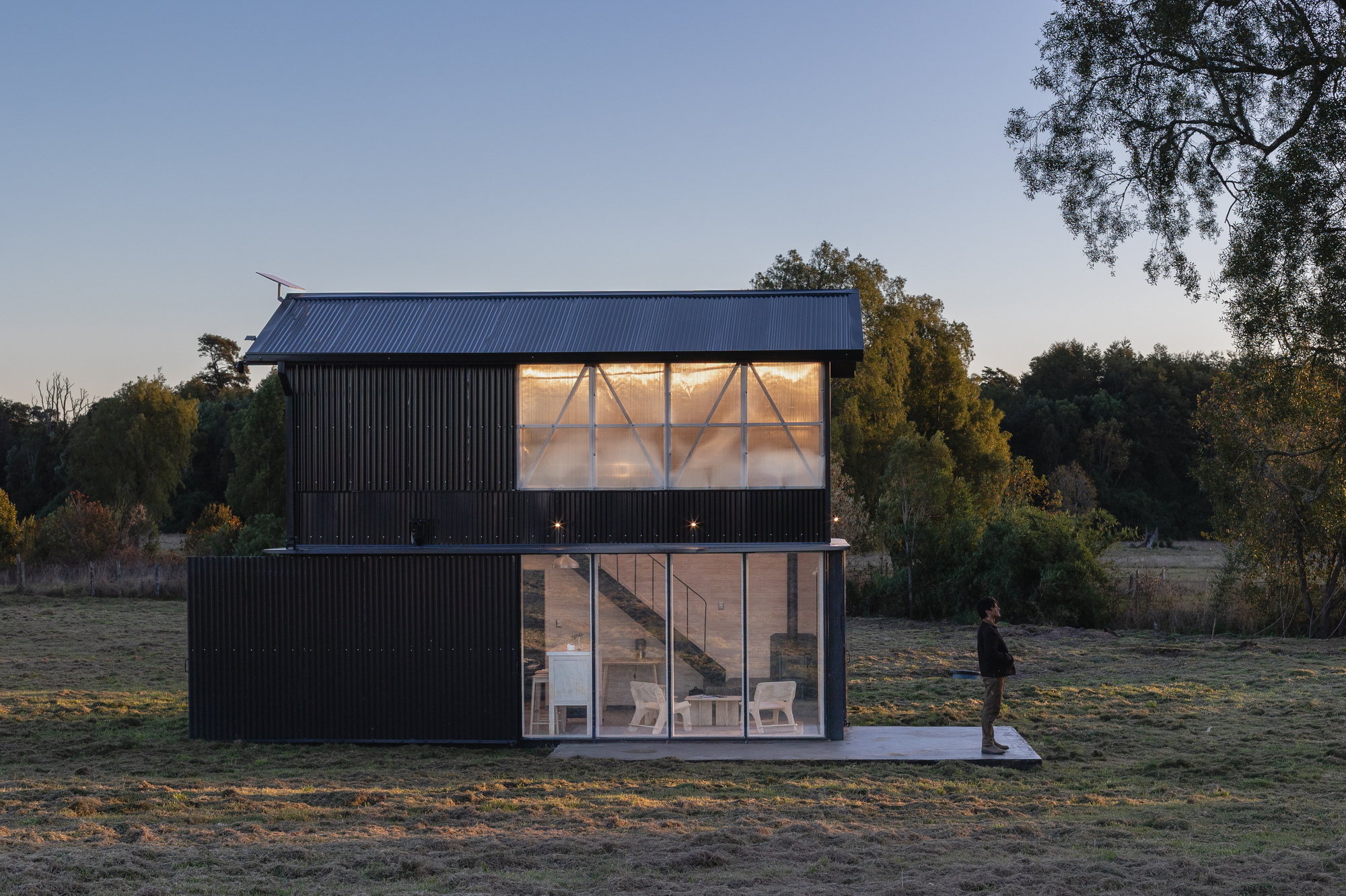 Side elevation of the black cabin with glowing translucent upper panels and glazed ground floor, a person standing on the concrete pad at dusk