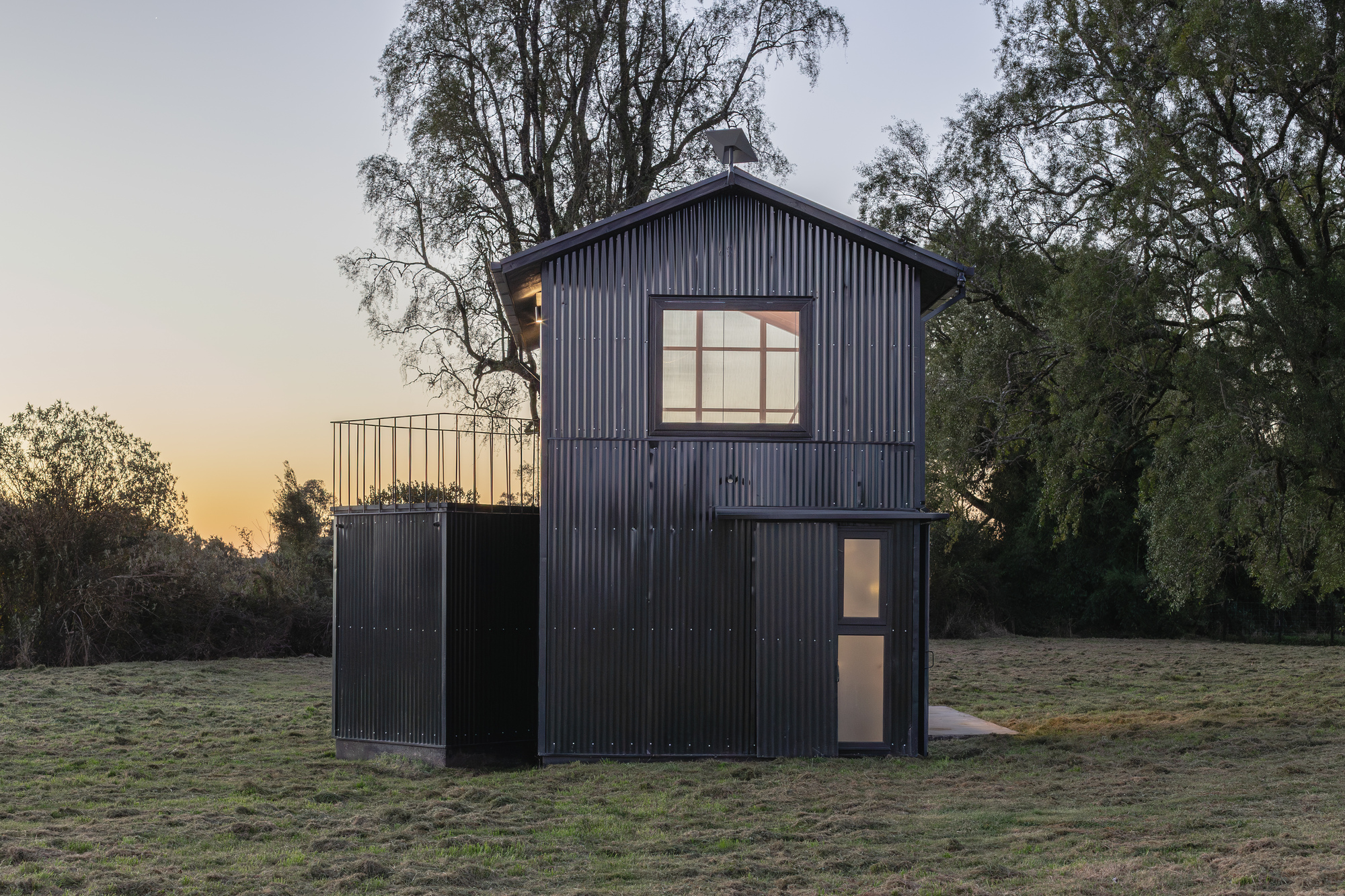Dark corrugated metal cabin with a gabled roof and lit upper window in a meadow at sunset