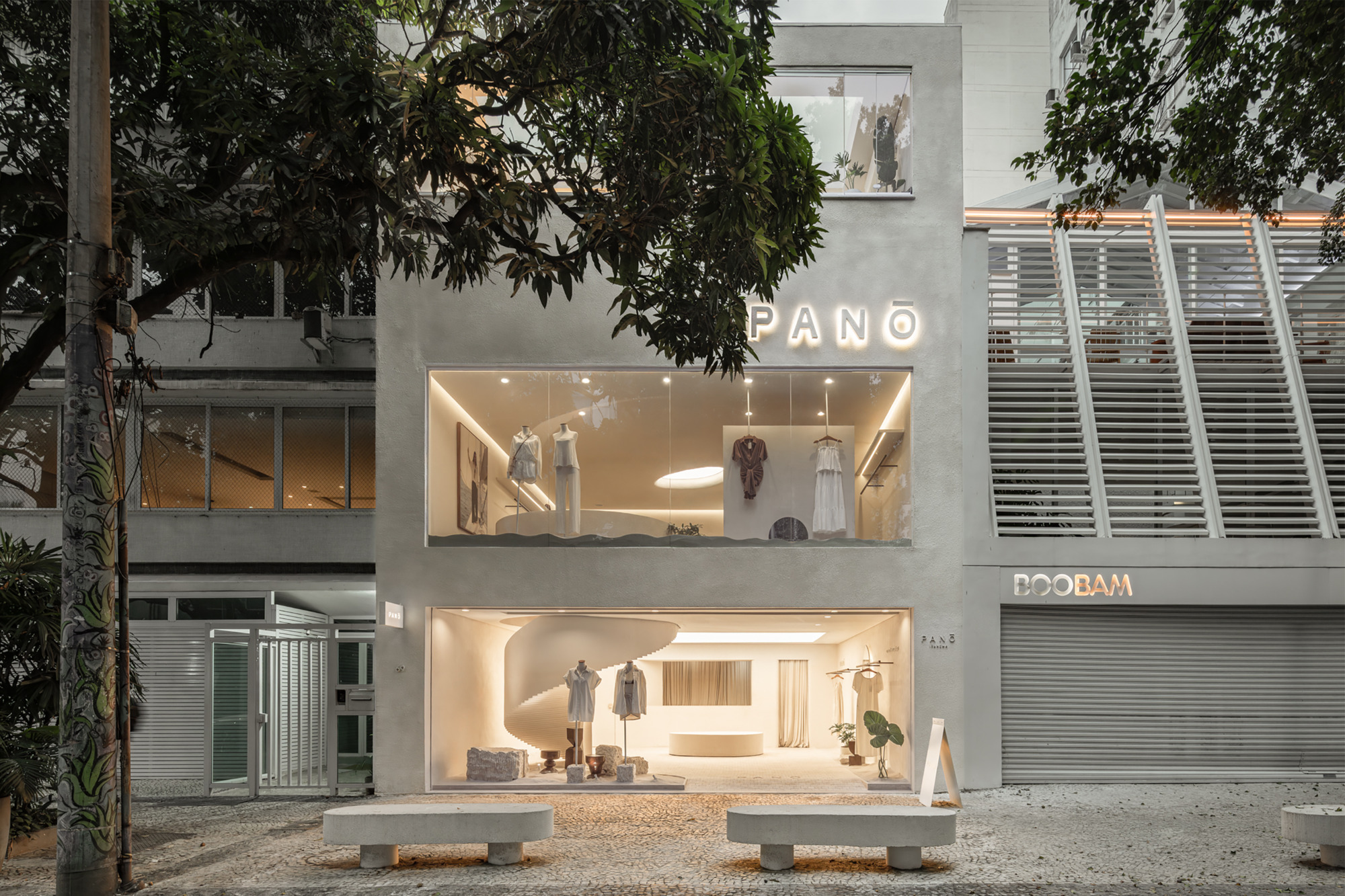 Gray plastered storefront with two glazed openings and illuminated lettering, stone benches in front and a mango tree overhead at dusk