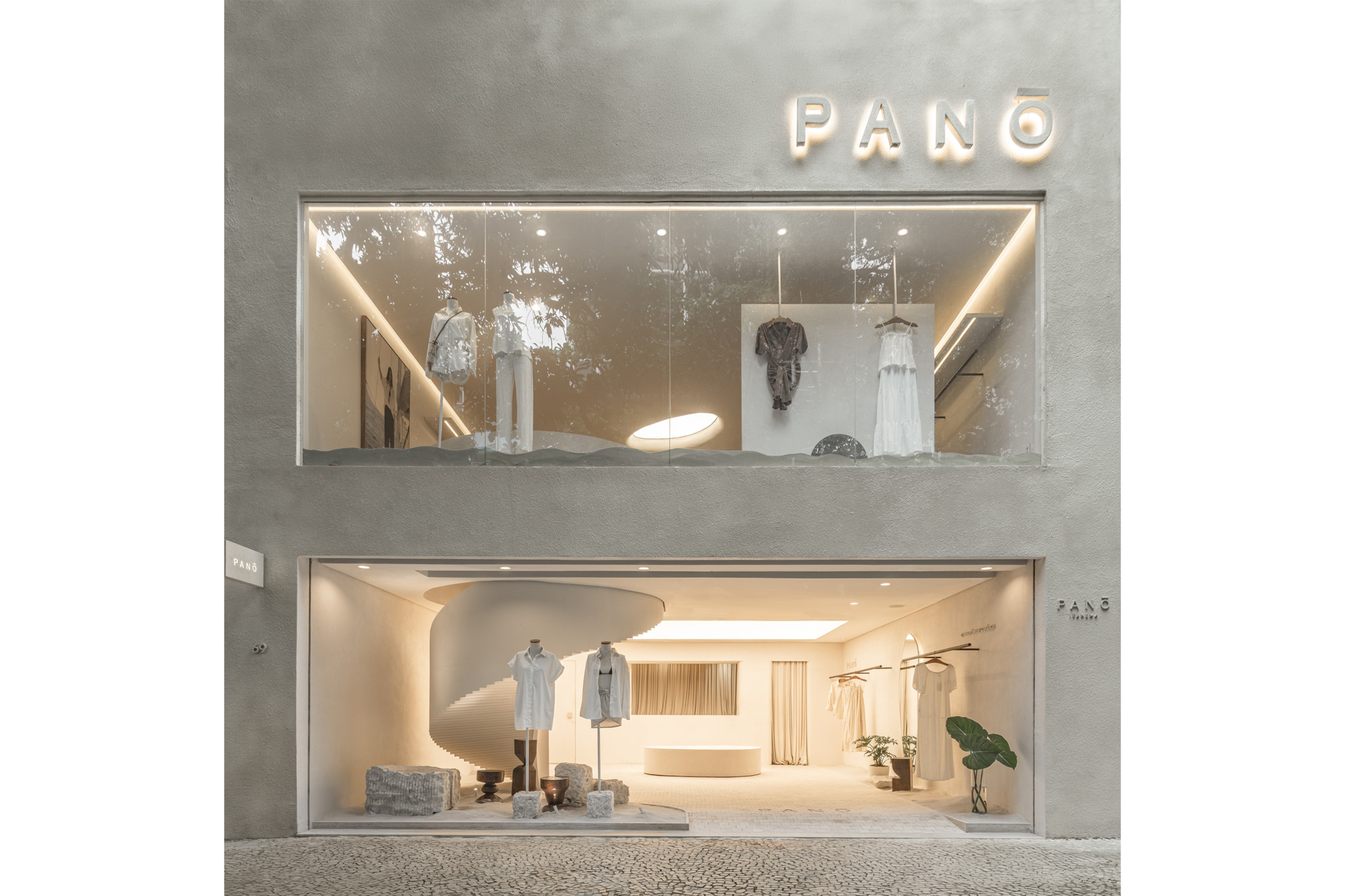 Frontal view of the two-level glazed shopfront showing a white spiral stair and hanging garments behind the glass, with tree reflections