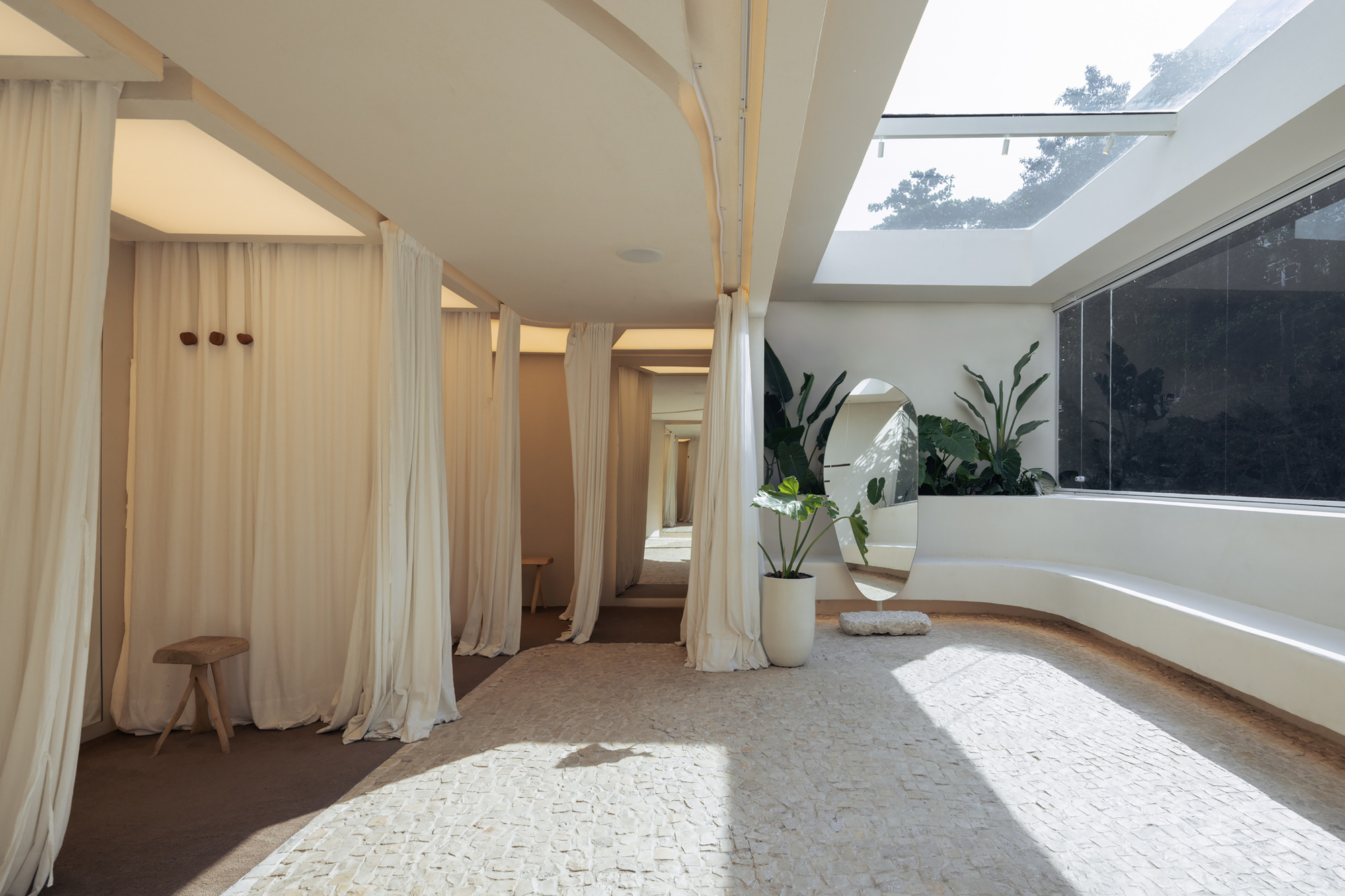 Fitting room corridor lined with cream curtains beside a curved built-in bench, a round mirror and a skylight with sun on the stone floor