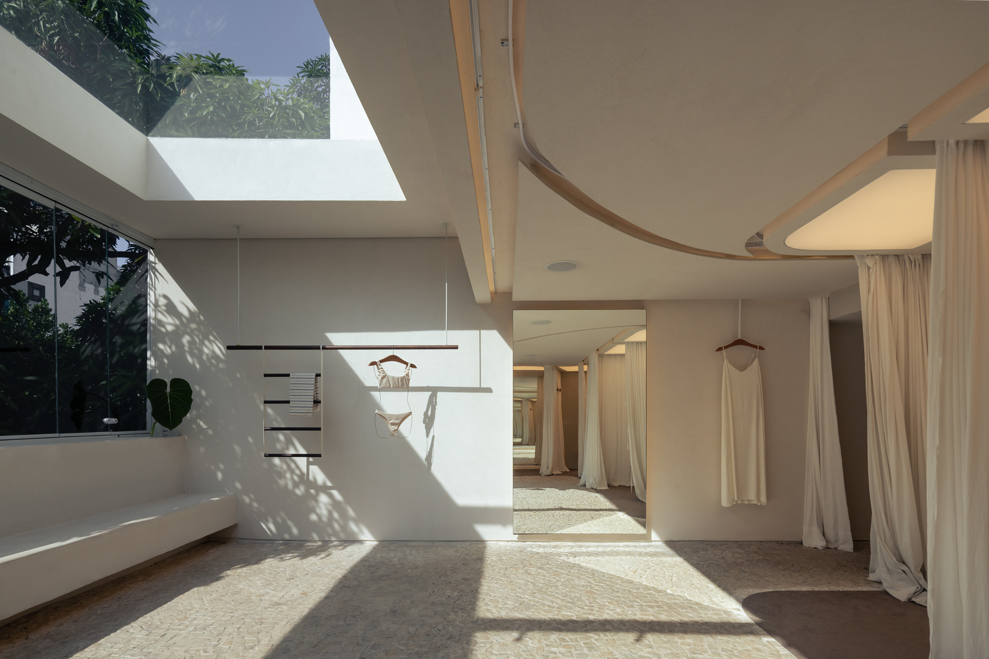 Interior with a skylight, curved ceiling edge and hanging garments, dappled leaf shadows falling across the pebble mosaic floor