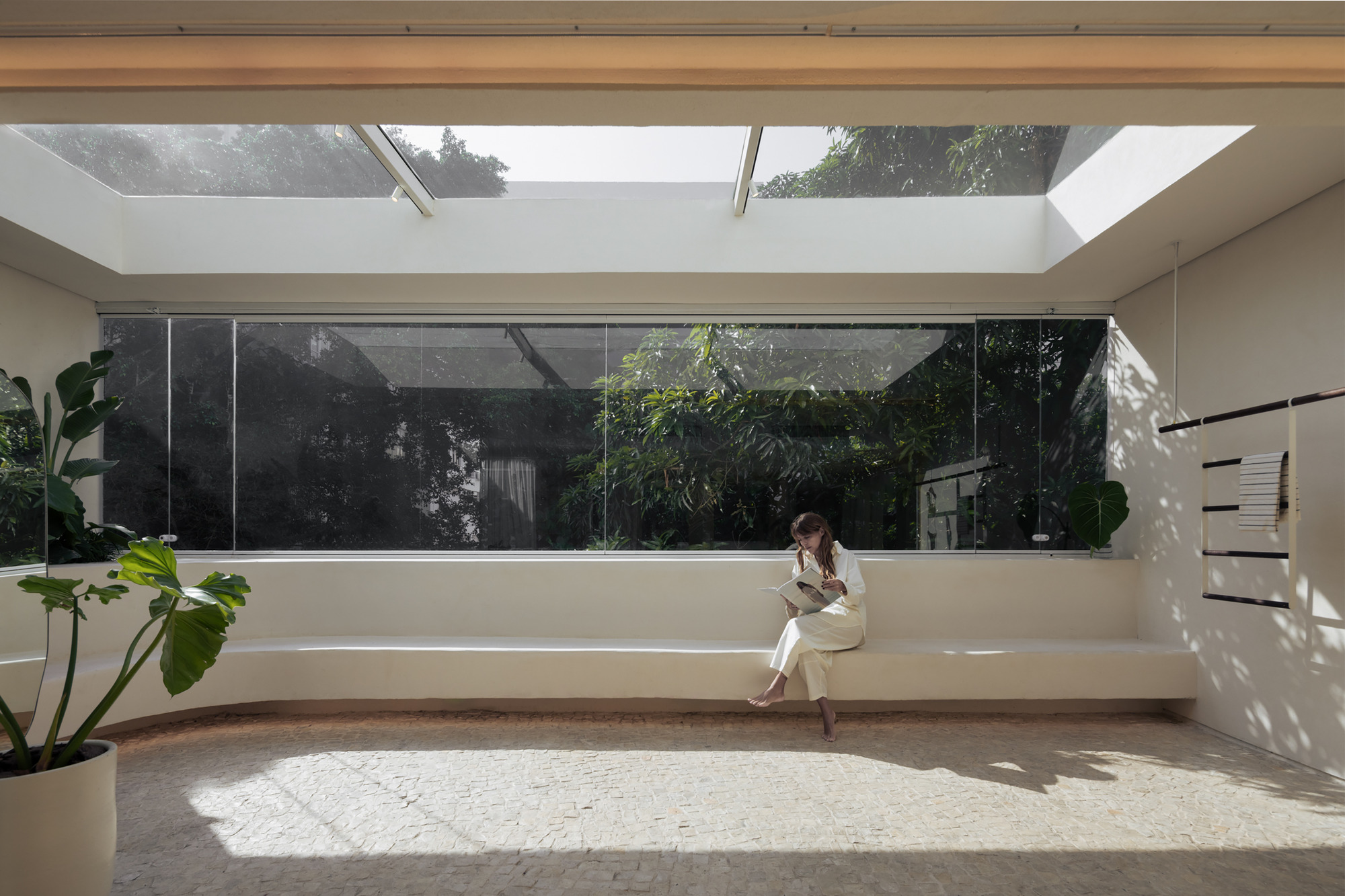Woman reading on a long curved plaster bench below a skylight, facing a glass wall of dense green foliage