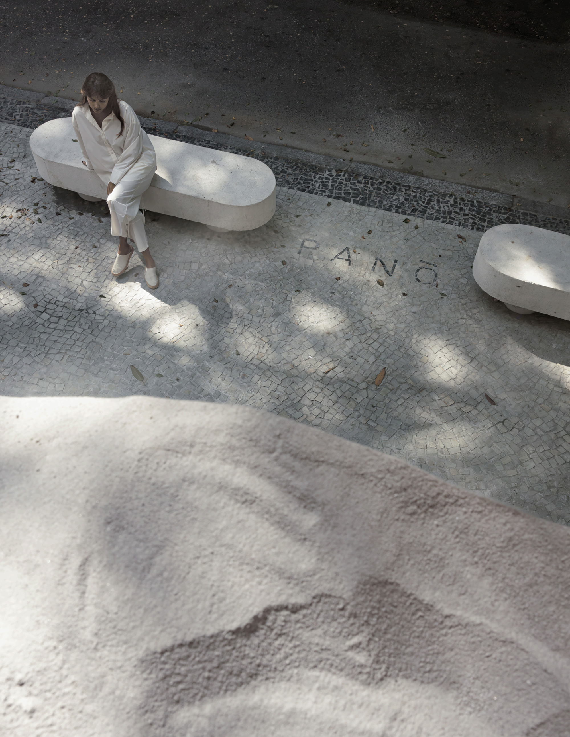 Overhead view of a seated woman on a stone bench over mosaic paving, with dappled tree shadow and a pale sand mound