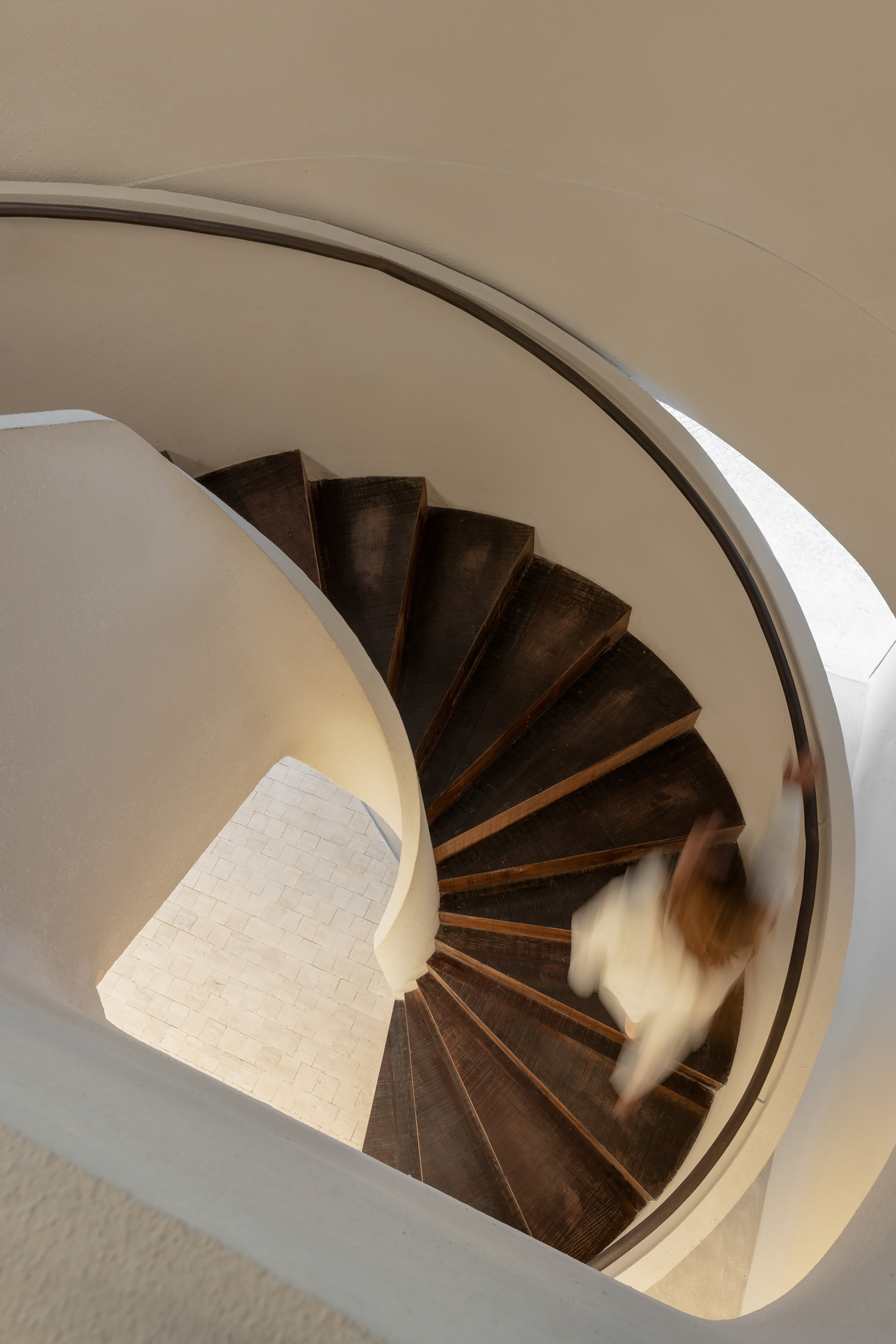 Looking down a spiral staircase with dark timber treads as a blurred figure in white descends past curved plaster walls