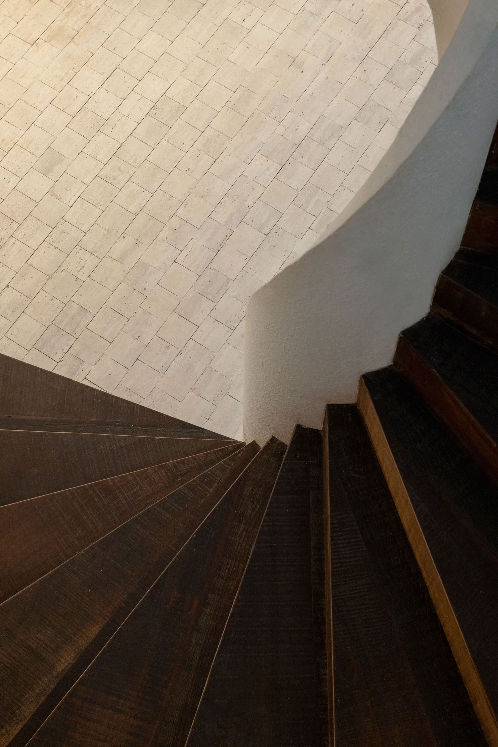 Top-down view of dark timber spiral stair treads curving beside a white plaster wall above pale stone paving in warm light