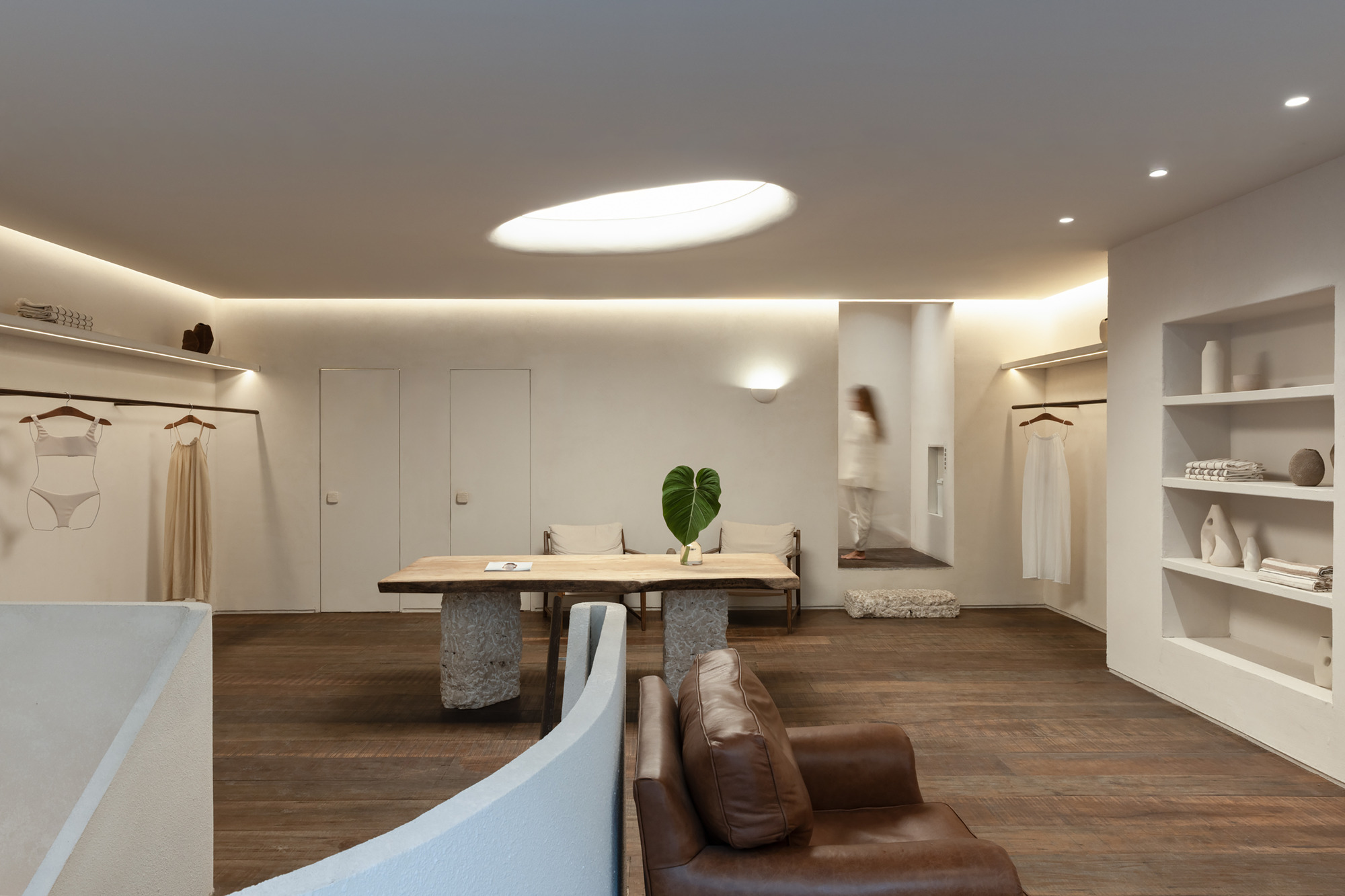Boutique interior with a live-edge timber table on stone bases, built-in shelves and a blurred figure walking past an oval skylight