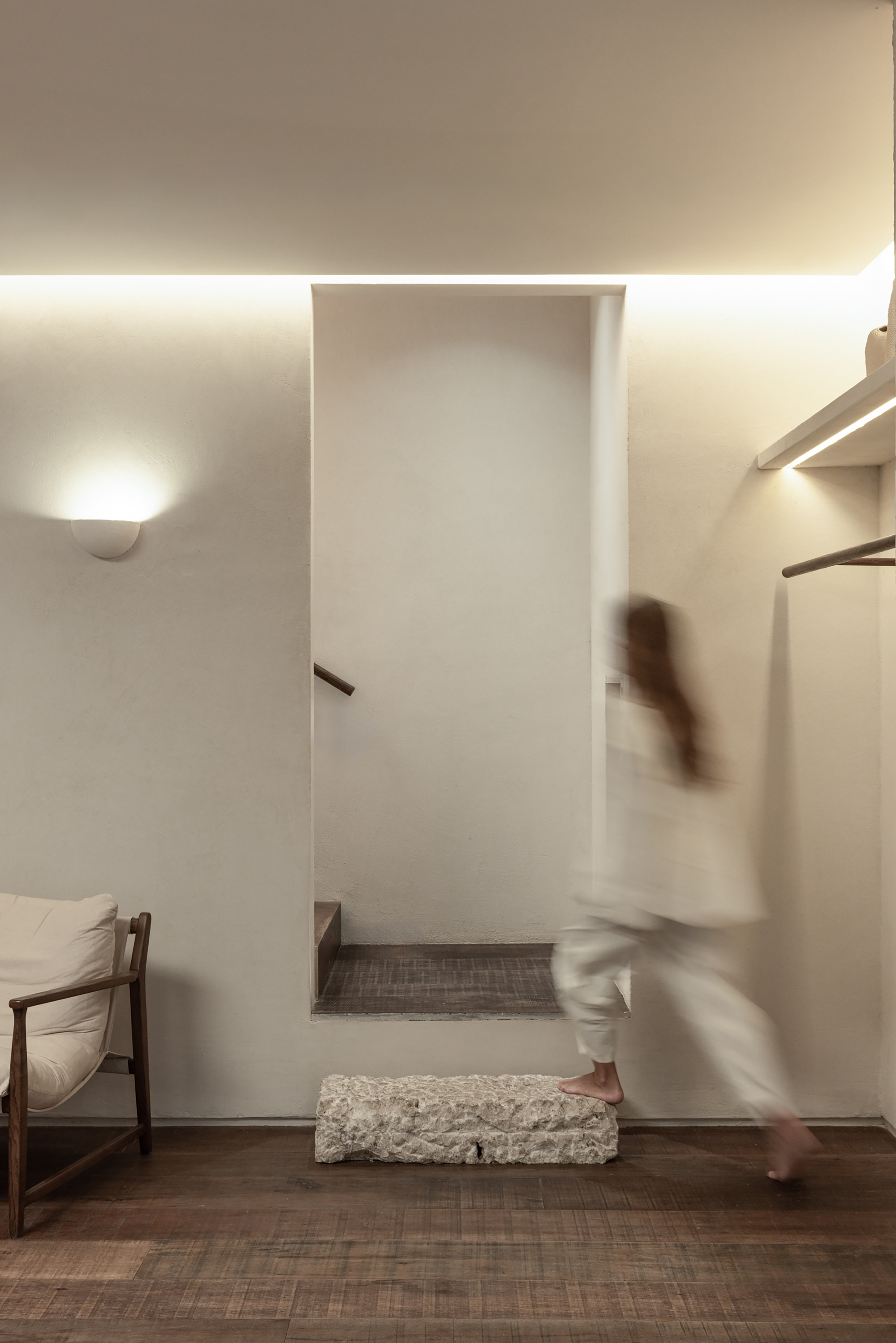 Plaster niche with a stair beyond and a rough stone block, as a blurred barefoot figure walks past under cove lighting
