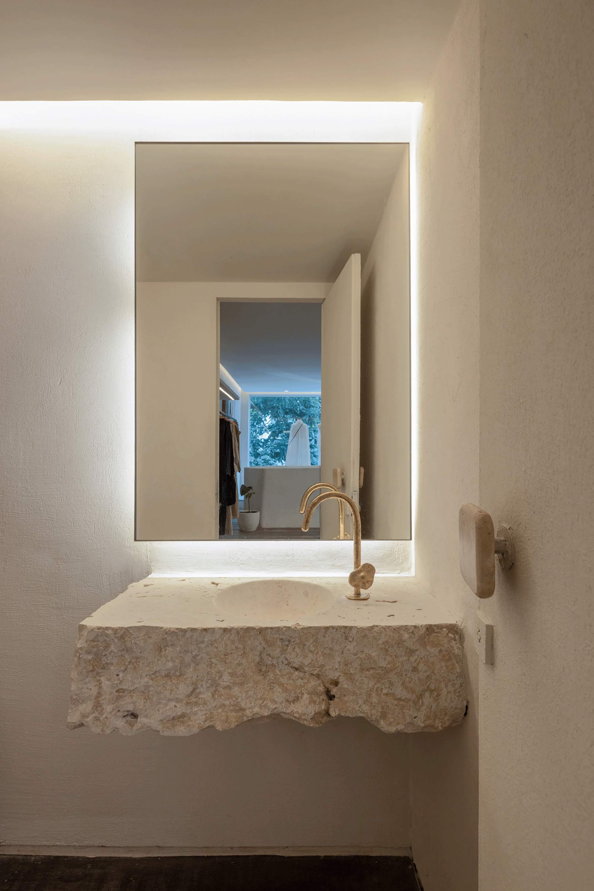 Rough-cut stone basin cantilevered below a backlit mirror reflecting a green tree seen through a distant window