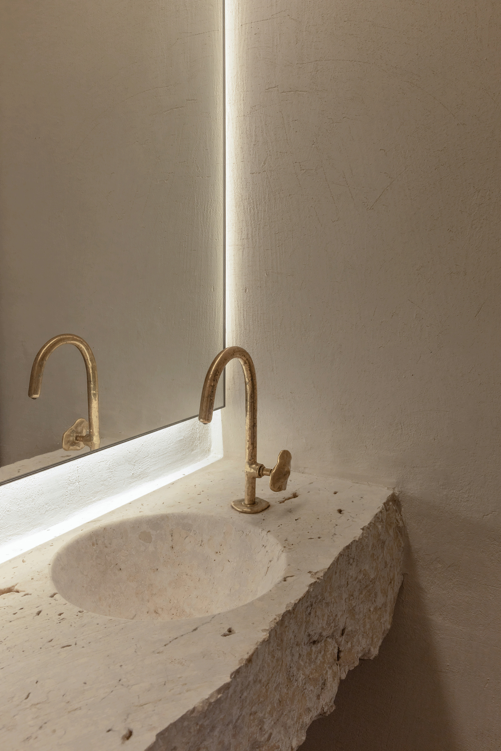 Close-up of a brass tap and carved stone basin beside a mirror edged with a thin line of light