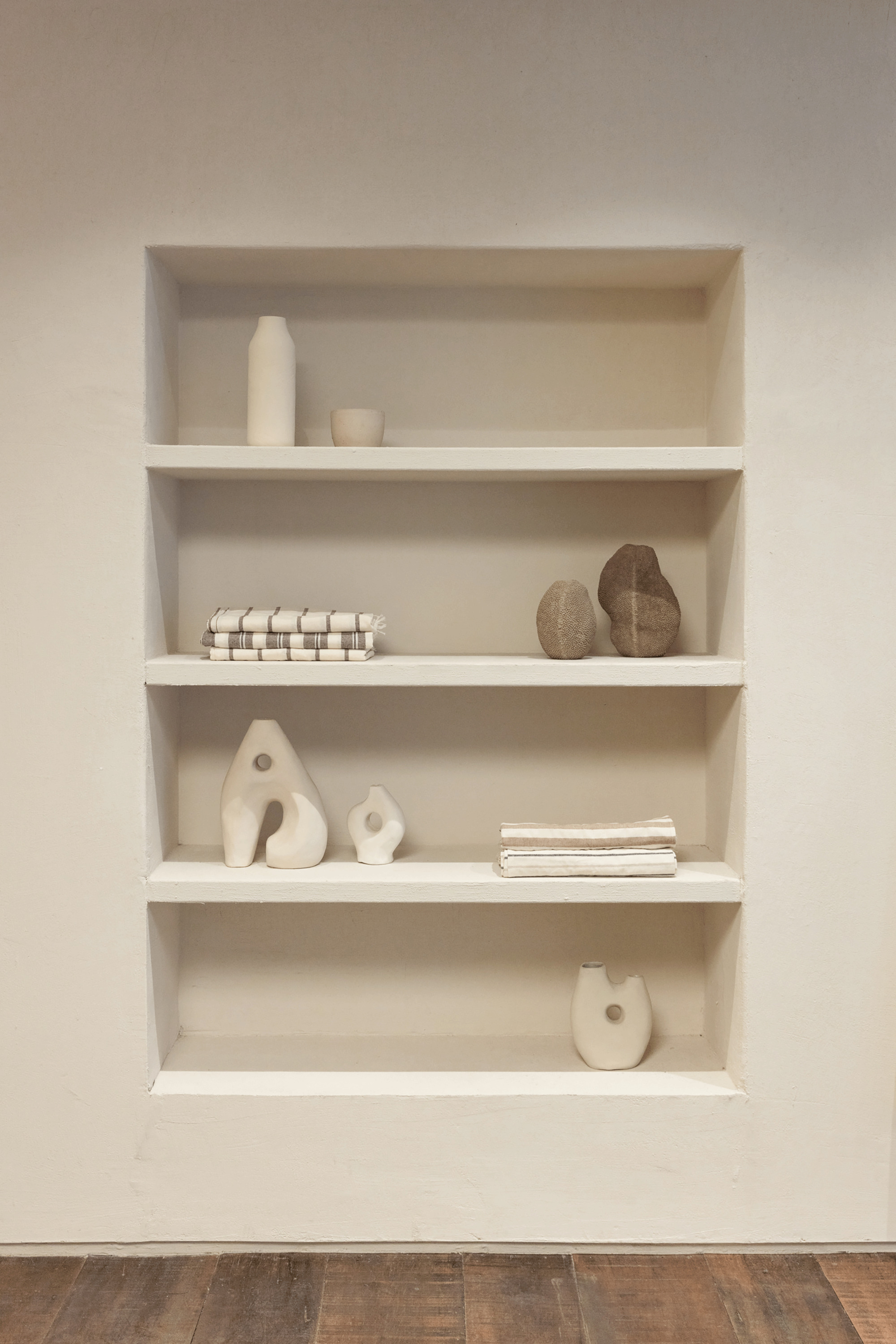 Recessed plaster shelving niche holding ceramic vessels, folded striped linens and two stones above a worn timber floor