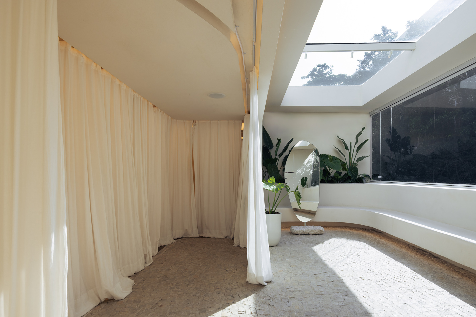 Sheer curtains enclosing a curved fitting area beside a skylight, a curved plaster bench and an oval mirror in afternoon sun