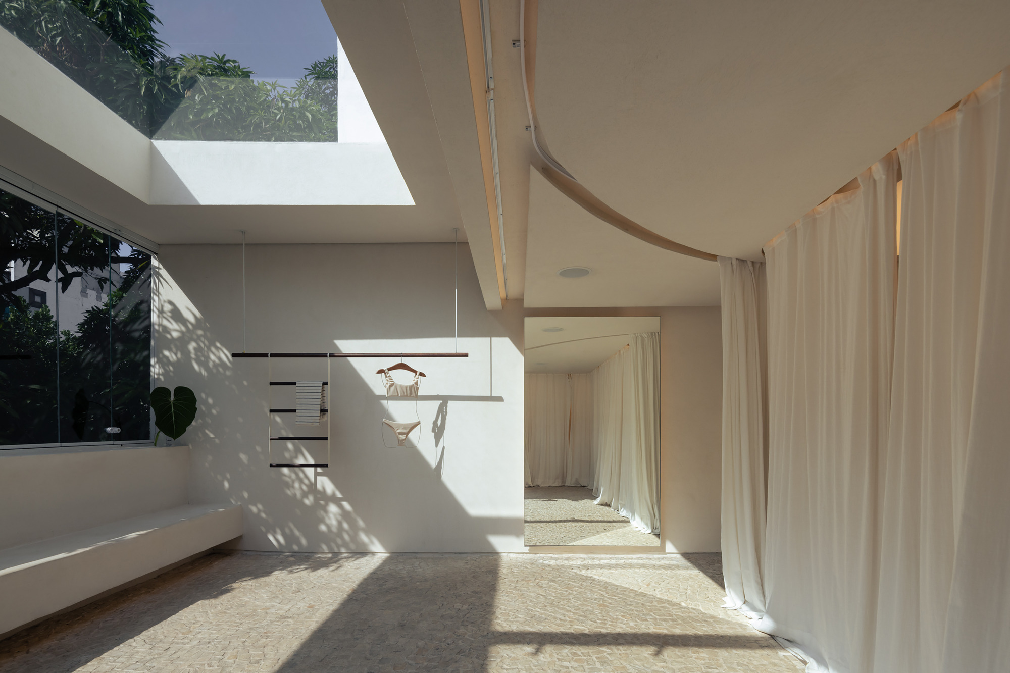 Skylight and glazed wall casting leaf shadows across a white wall with a hanging rail, beside draped curtains and a mirror