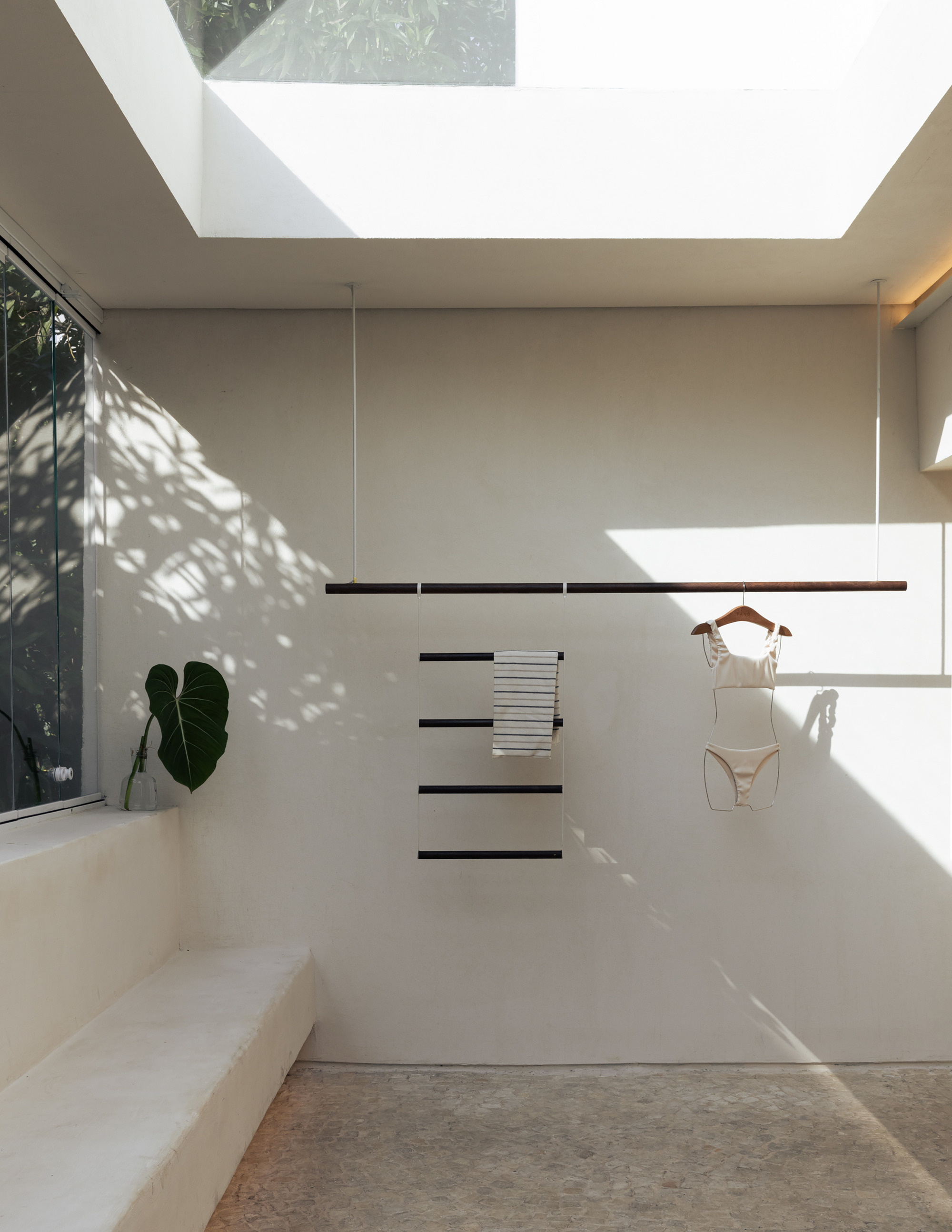 Suspended timber rail with a striped towel and swimsuit on a white wall, dappled with leaf shadows beneath a skylight