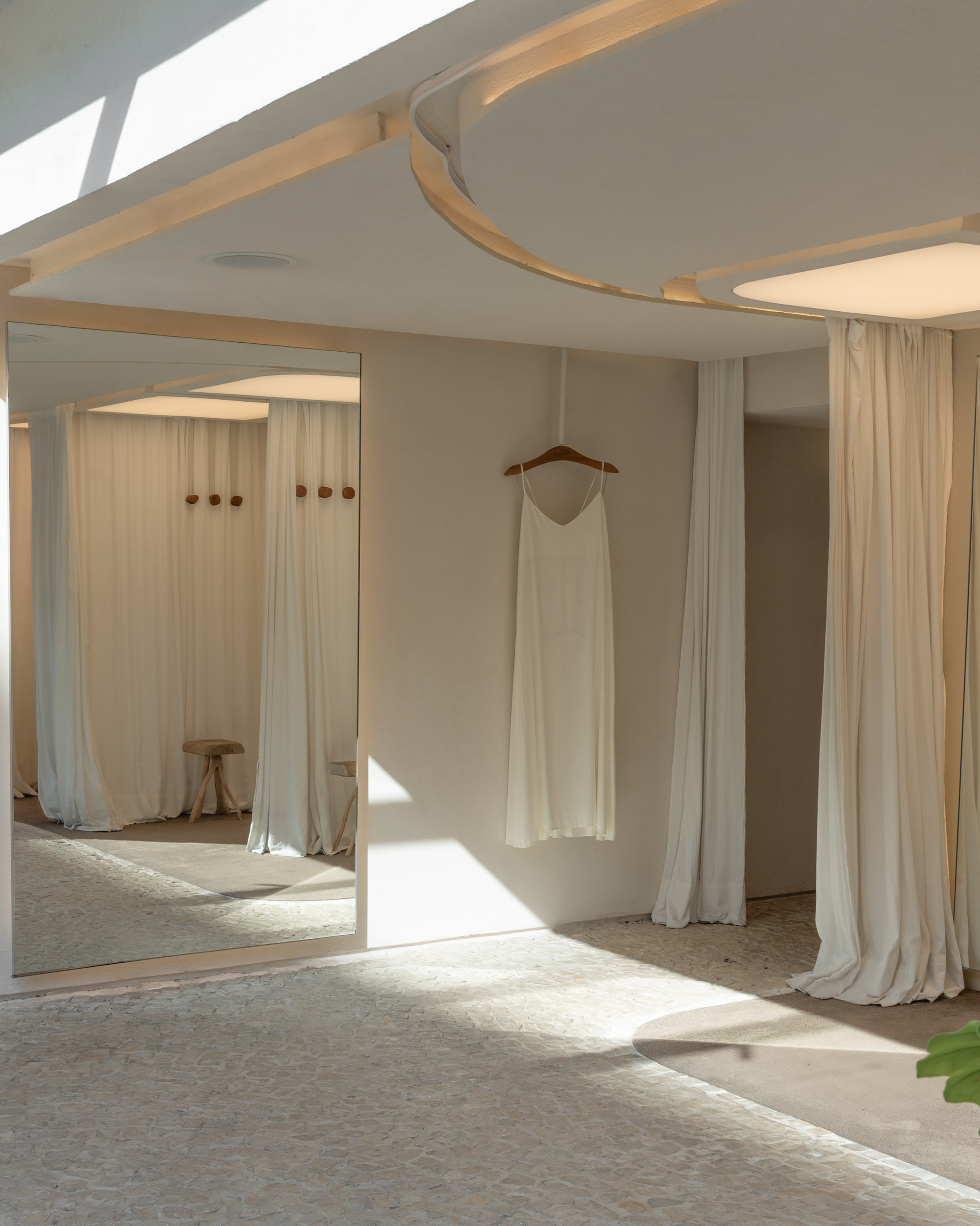 Full-height mirror reflecting curtained fitting rooms, with a slip on a hanger beside it and a curved ceiling edge glowing above