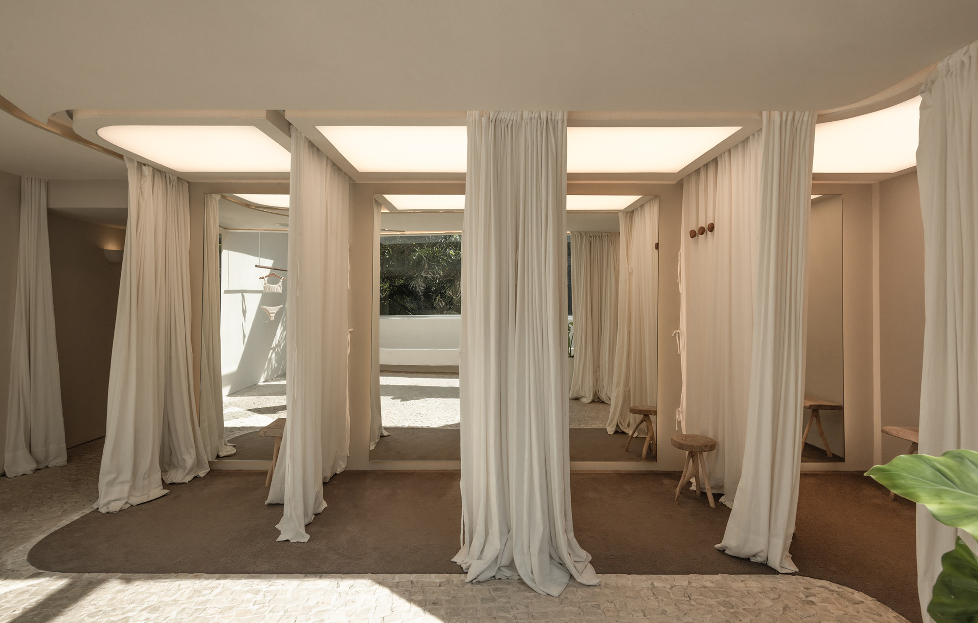 Row of curtained fitting cubicles with wooden stools and backlit ceiling panels, opening toward a planted courtyard in daylight