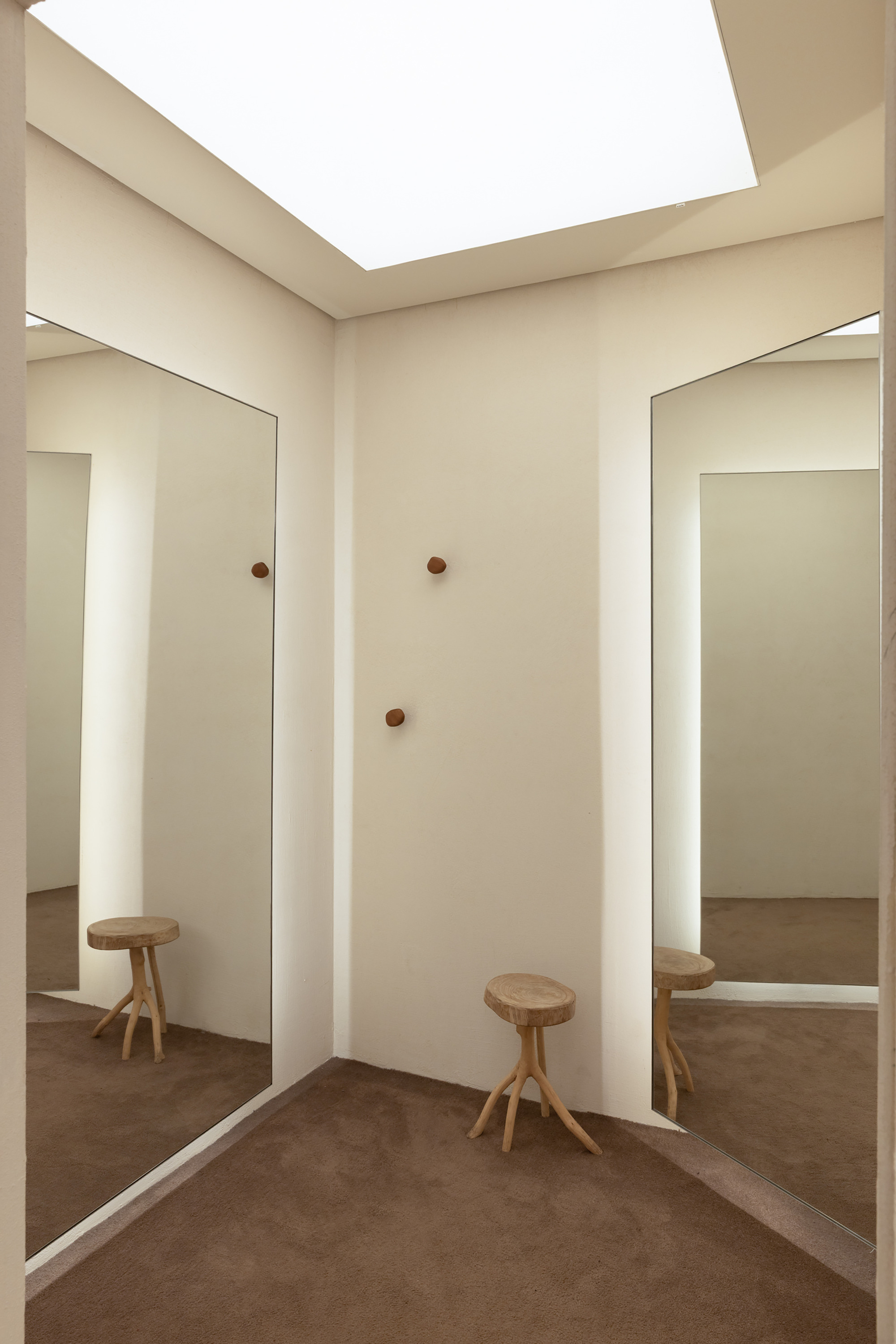 Fitting room corner with a skylight, full-height mirrors, wall pegs and two branch-legged wooden stools on brown carpet in soft daylight