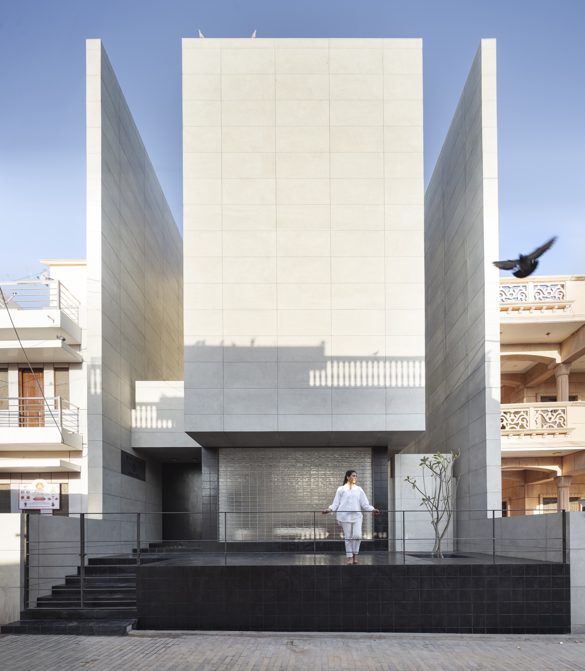 Front facade of pale stone panels with a black tiled plinth, a woman standing on the terrace and a bird flying past at dusk