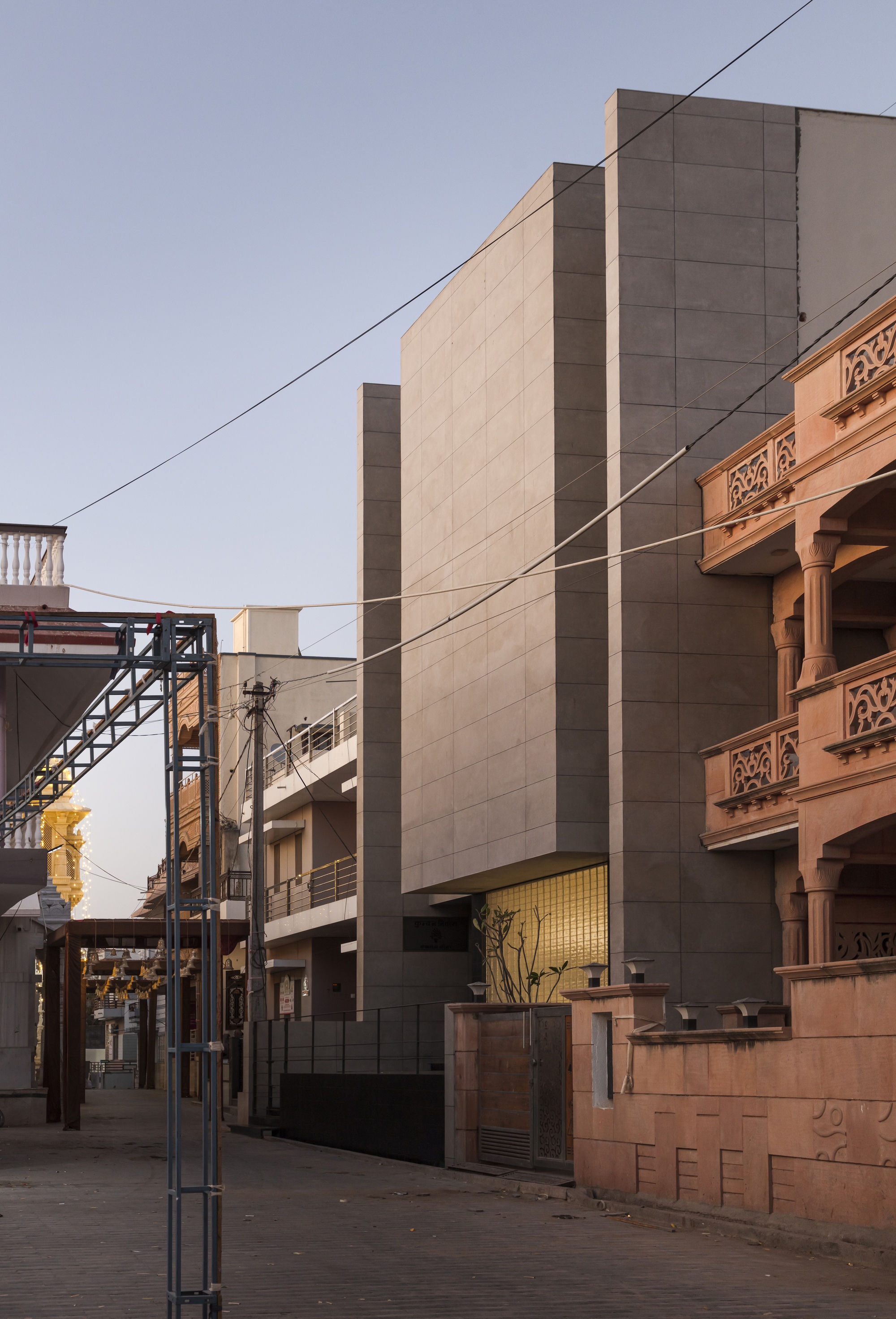 Side street view of the grey panelled volume rising beside a carved pink sandstone neighbor, with overhead wires in evening light