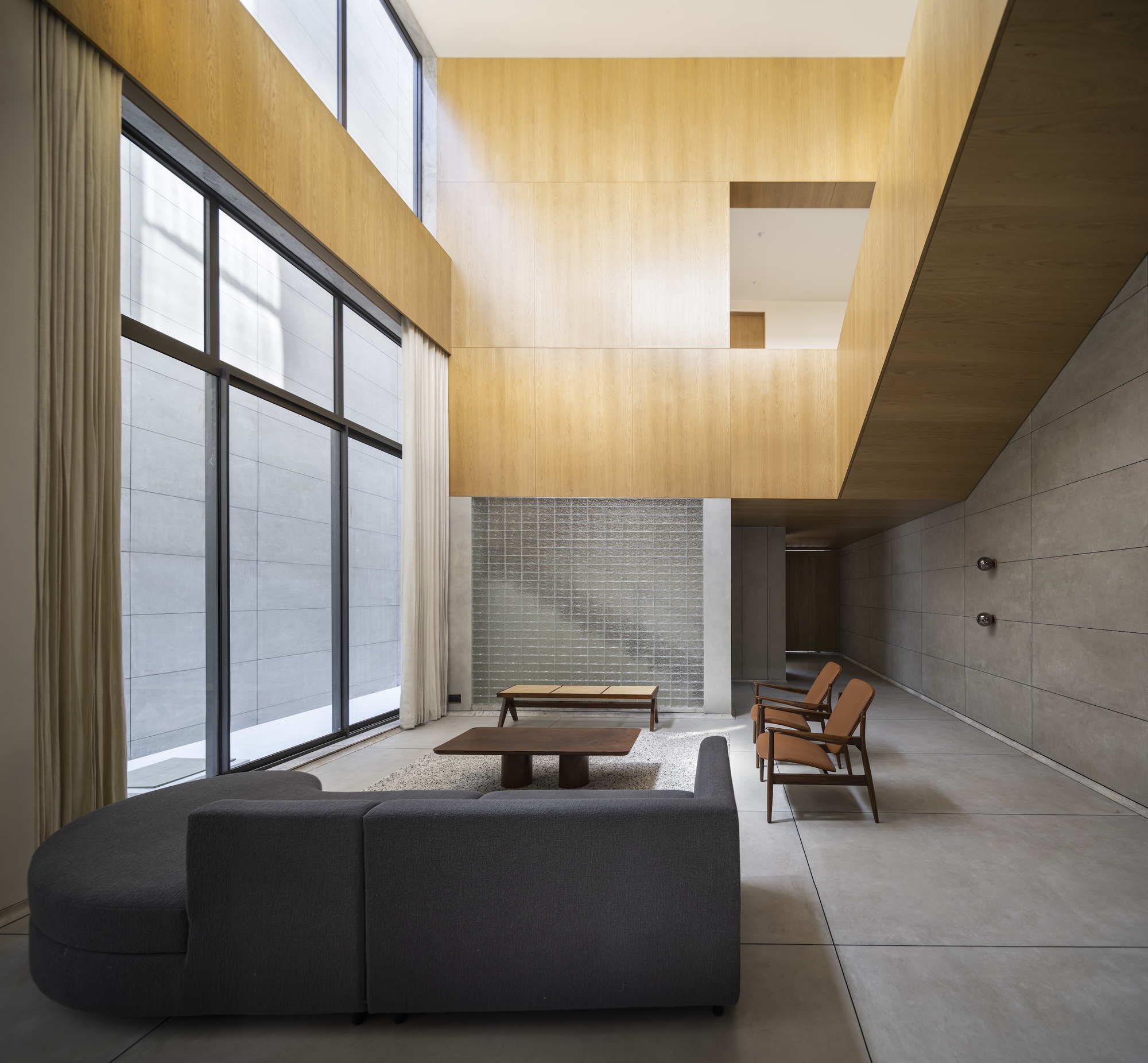 Double-height living room with oak paneling, glass block wall, grey sofa and leather armchairs beside a tall window with linen curtains