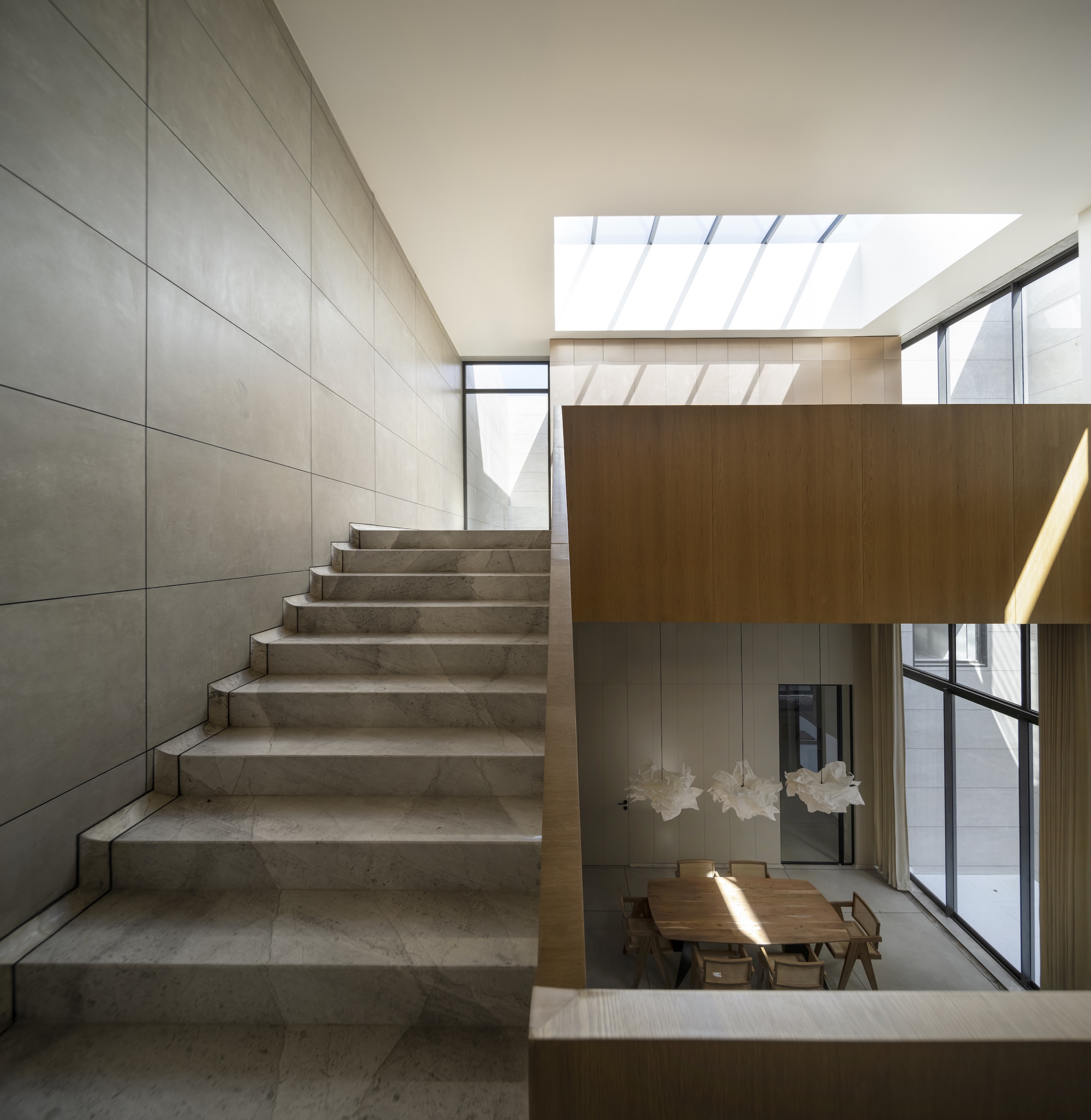 Stone staircase beside a panelled wall, with an oak balustrade overlooking a dining table under a skylight and paper pendant lamps