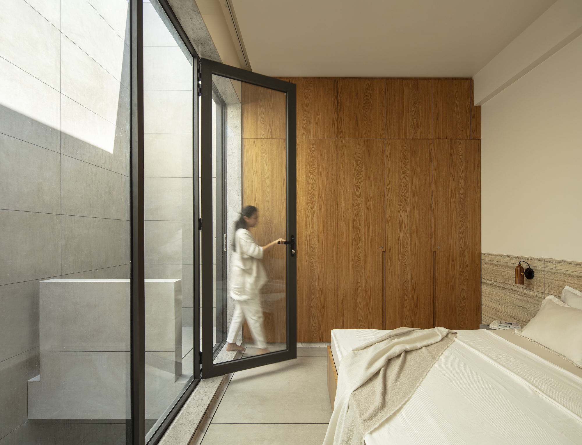 Bedroom with oak wardrobe wall and an open black-framed glass door, a blurred woman walking past a light well