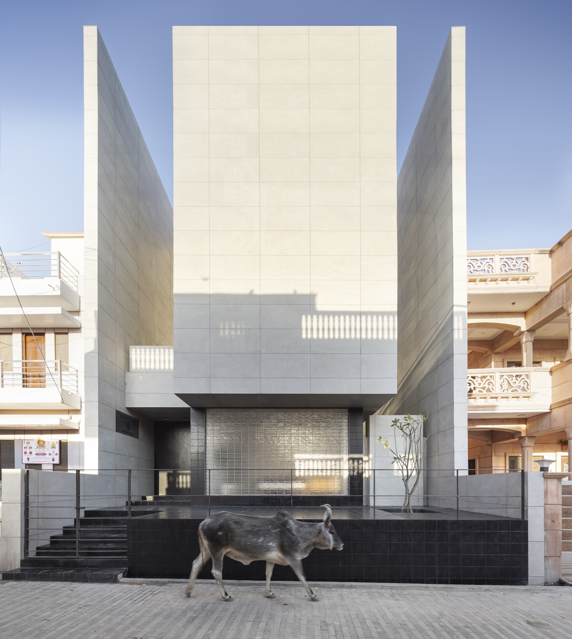 Street facade of pale stone panel walls flanking a glass block entrance, with a cow walking past in clear daylight