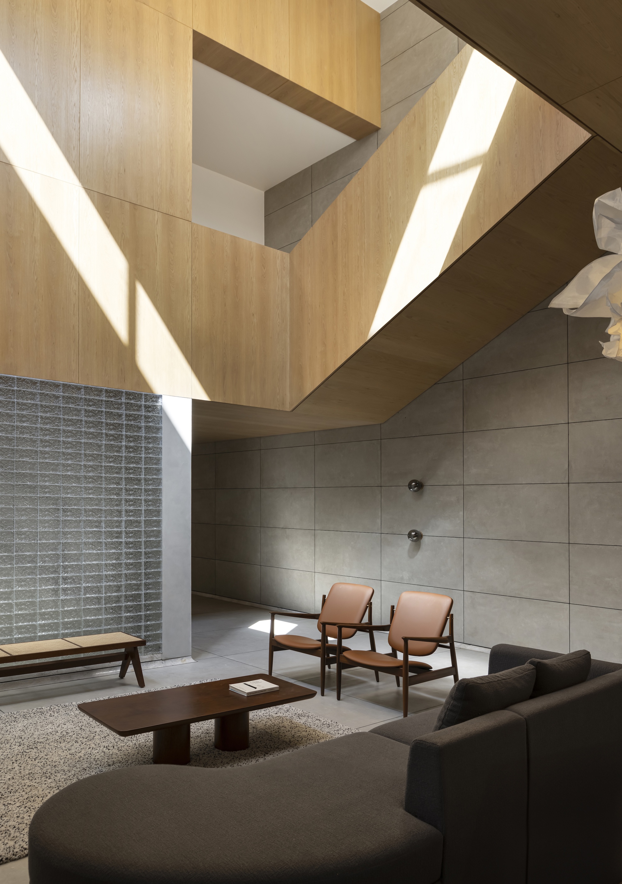 Double height living room with oak panelled mezzanine, concrete panel wall and shafts of sunlight falling across the sofa and chairs