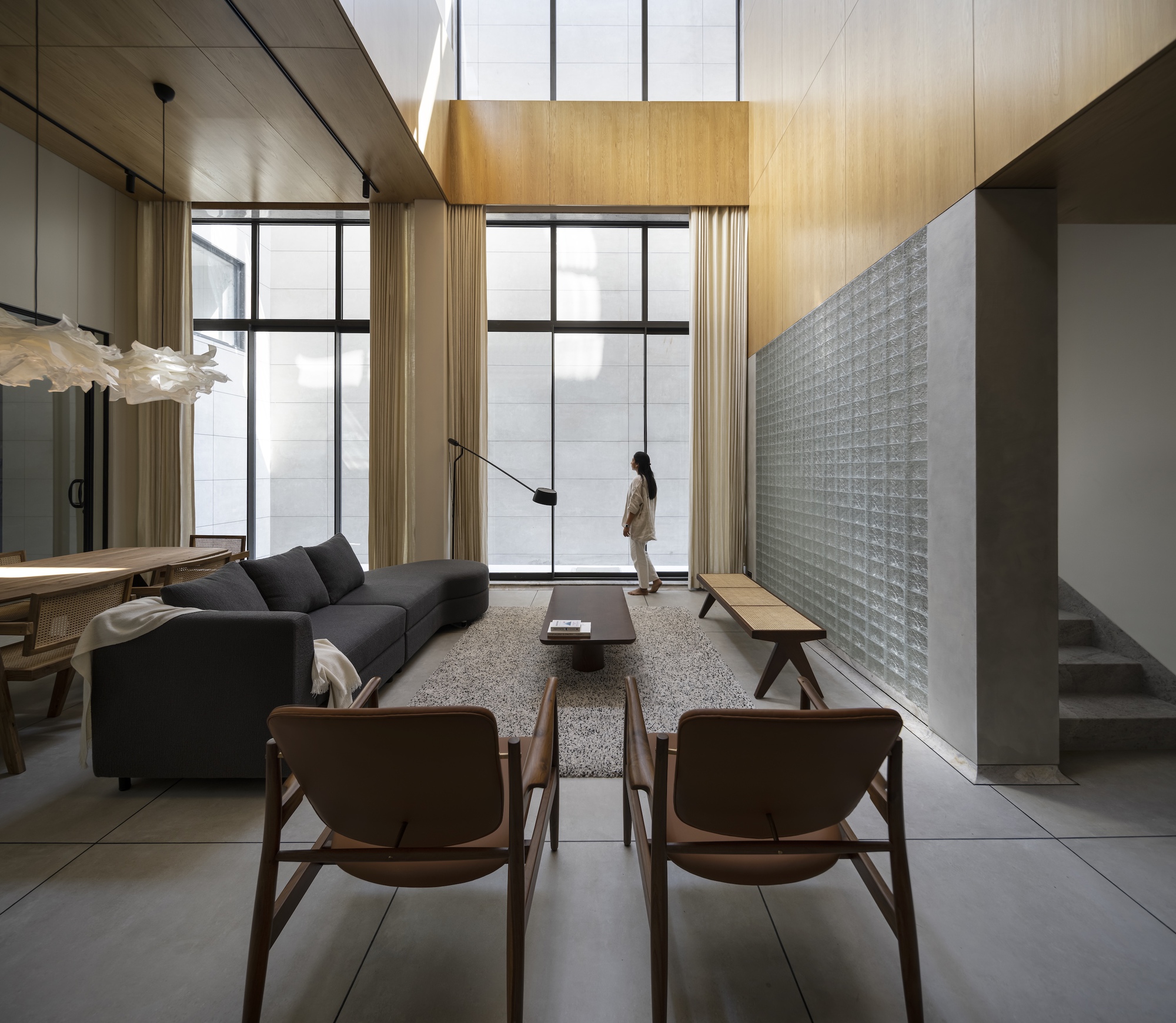 Living room seen from the chairs toward tall windows, with a woman standing at the glass beside a glass block wall