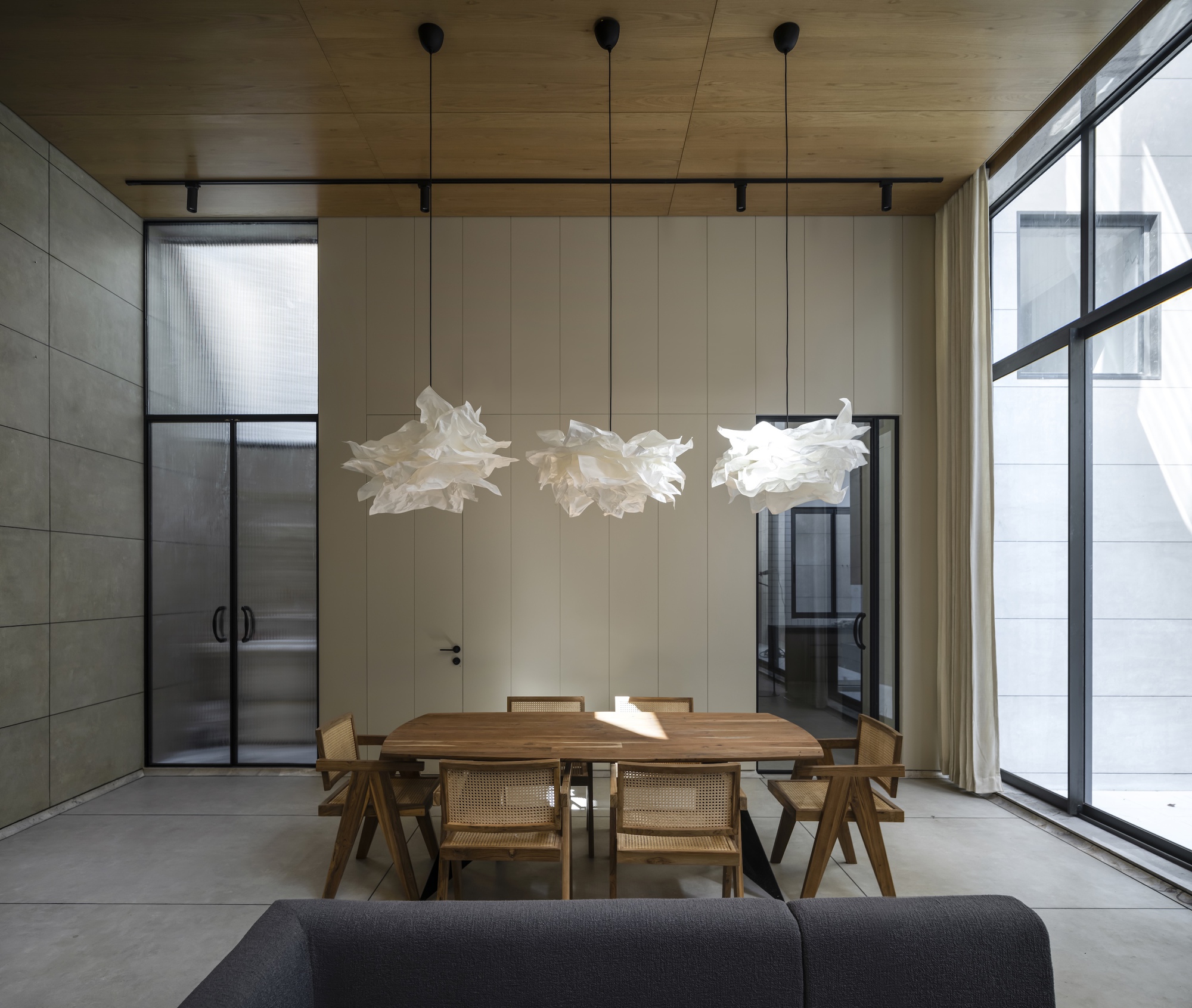 Dining table with cane chairs under three paper pendant lamps, set against a pale panelled wall and tall curtained windows