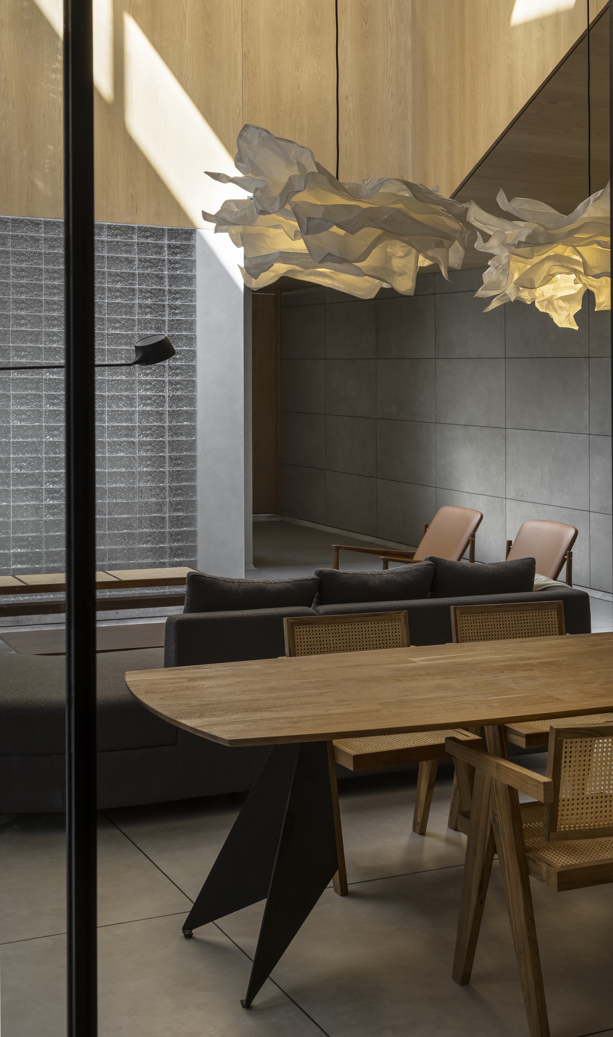 Dining table with a black angular base and cane chairs, with paper pendant lamps and a glass block wall behind in raking sunlight