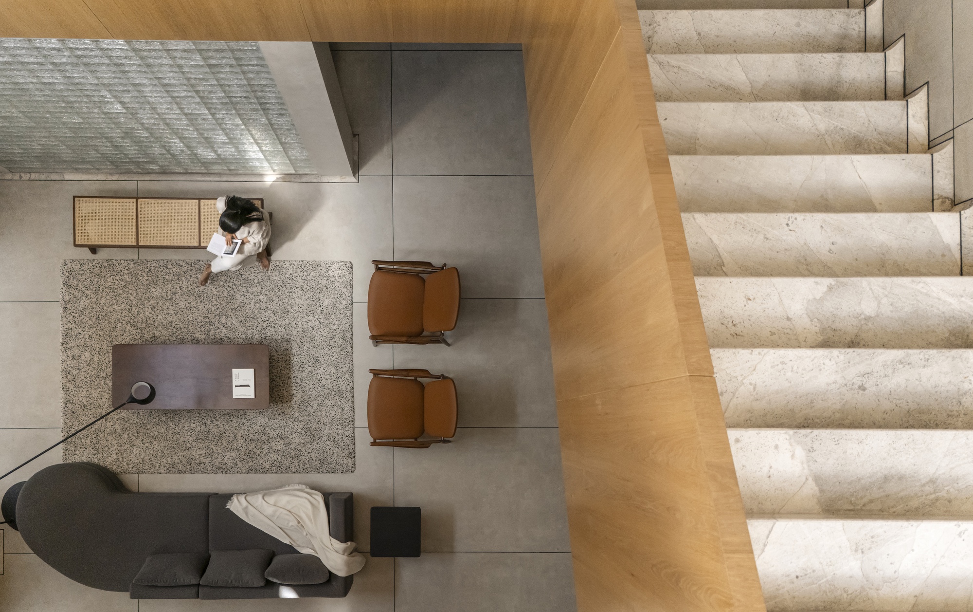Top-down view of a living area with rug, leather armchairs and sofa beside a marble stair, a woman reading at the edge