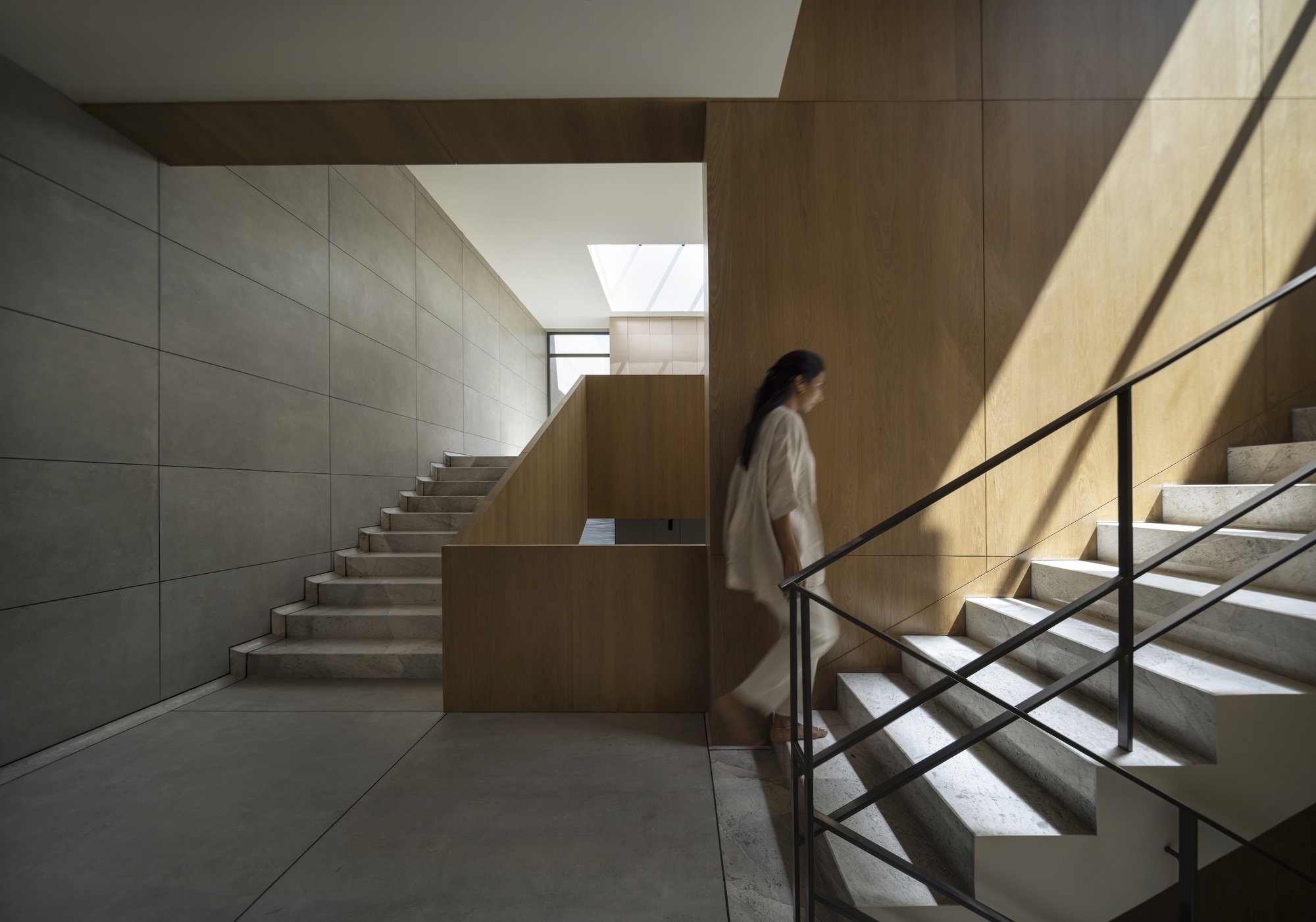 Marble stair with a black steel handrail and oak paneling, a woman walking past as sunlight cuts across the wall