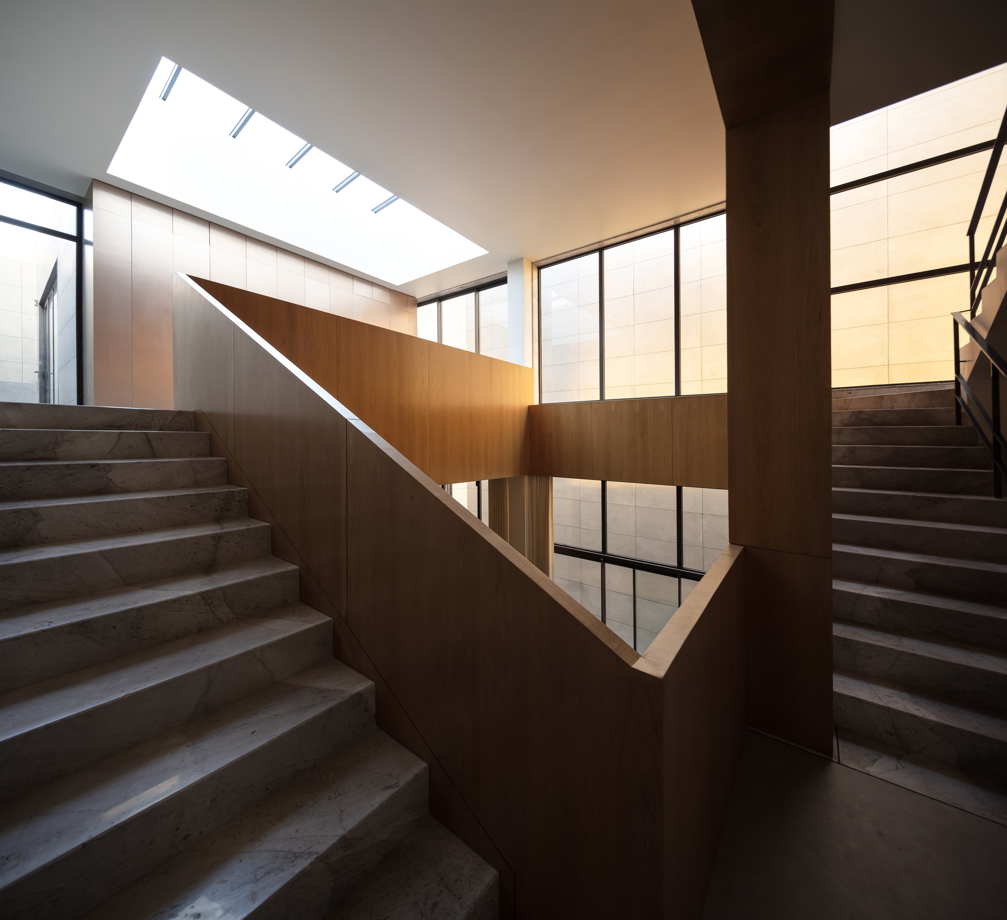 Wide-angle view of timber-clad stair landings and marble treads beneath a skylight, with tall windows glowing in warm late light