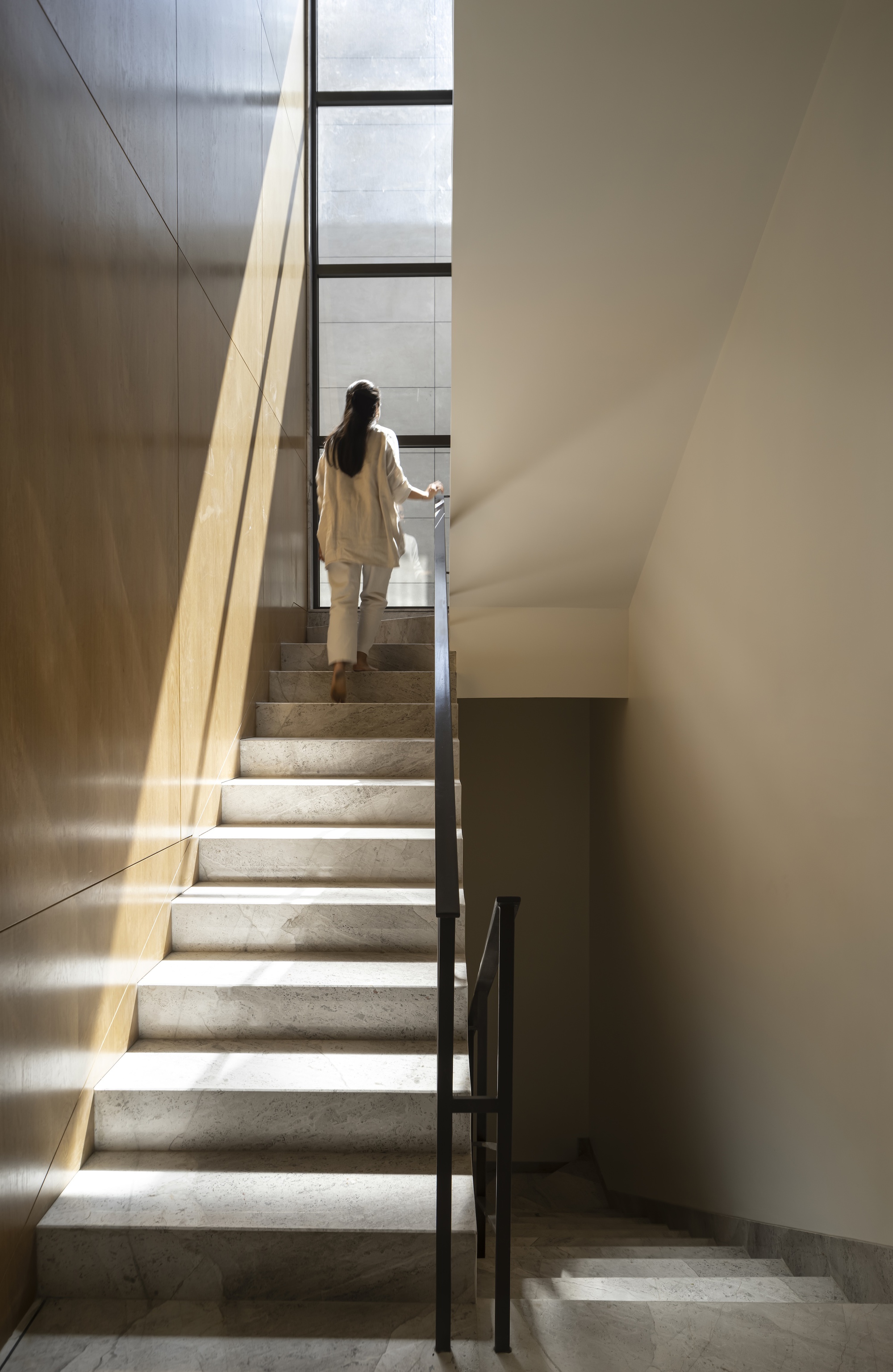 Woman climbing a narrow marble stair beside an oak panel wall, with a tall window ahead and a shaft of sunlight
