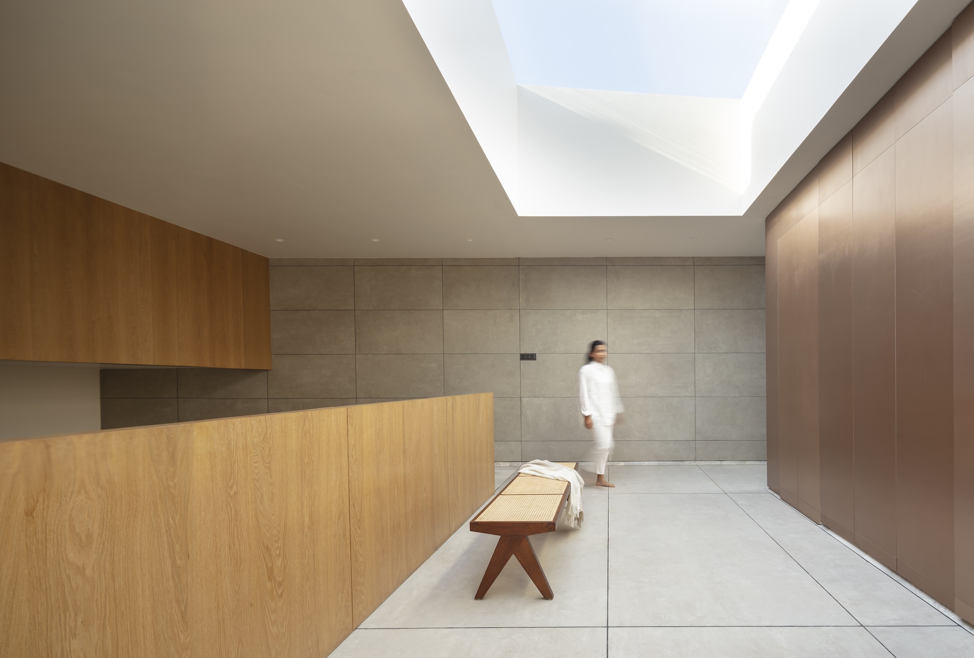 Top-lit landing with a cane bench, oak balustrade and concrete tile wall, a woman in white passing under the skylight