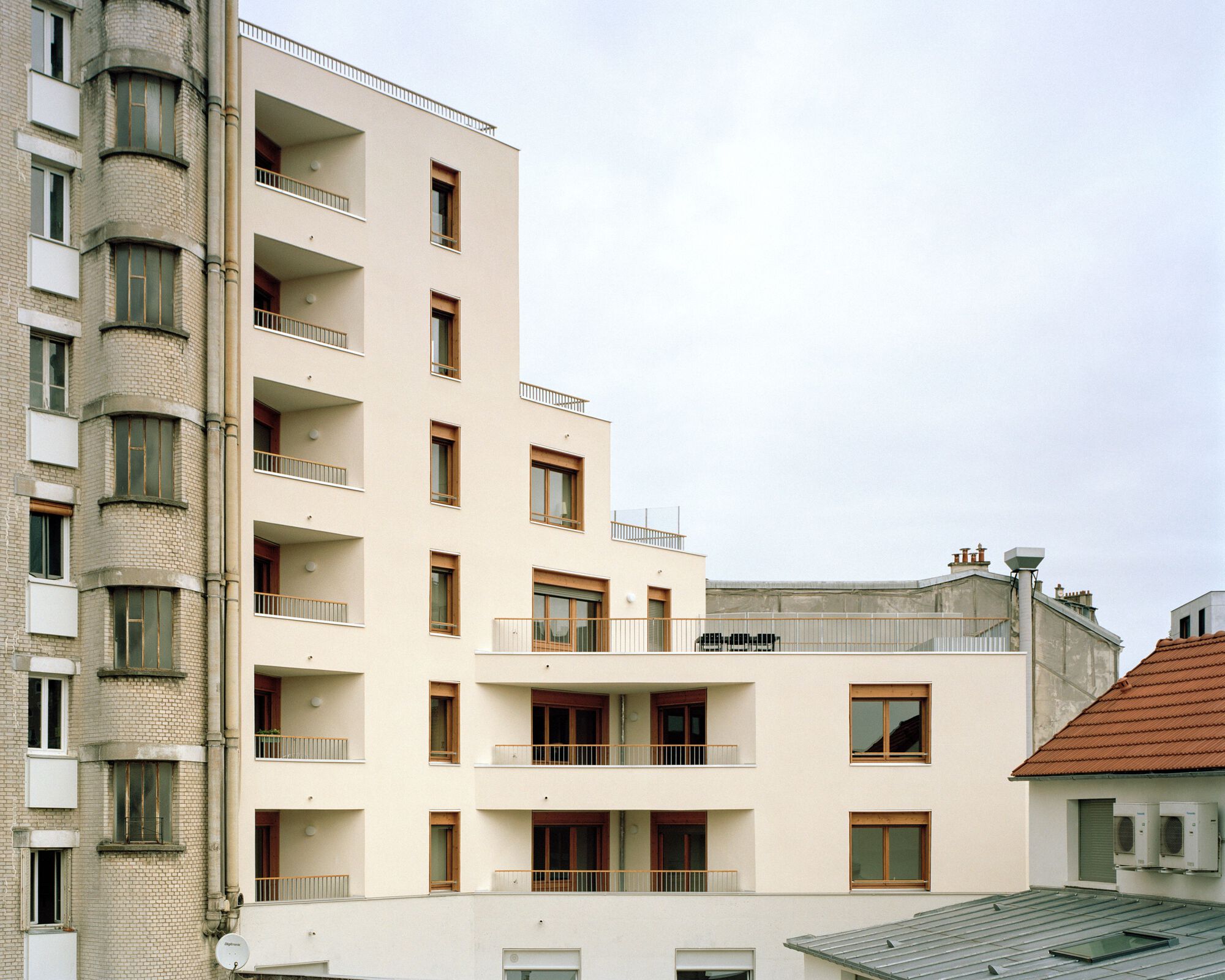 Stepped rear facade of a cream rendered block with timber window frames and recessed balconies beside a brick turret under overcast sky