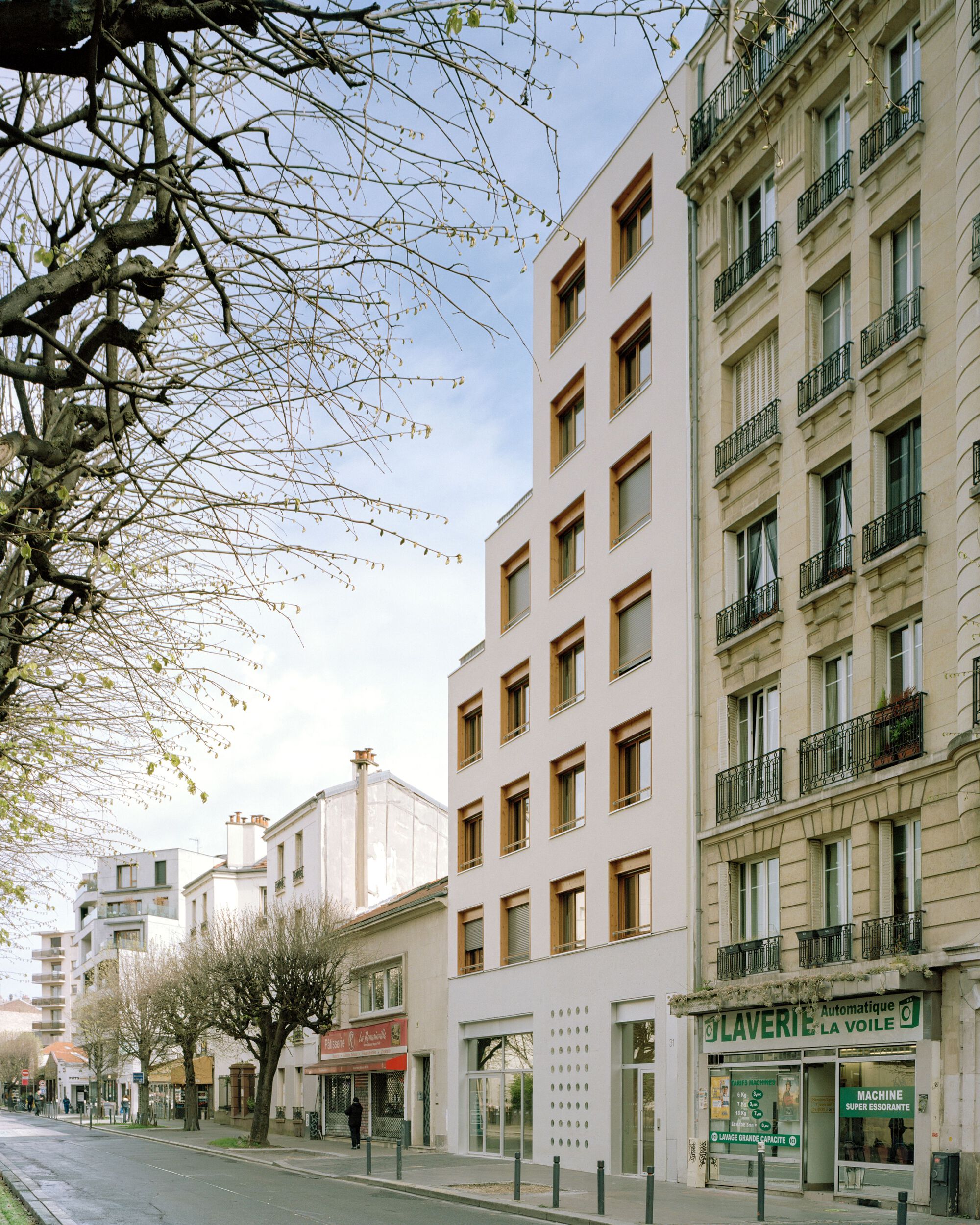 Street view of a narrow white building with timber-framed windows wedged beside a stone apartment block, bare tree branches in the foreground