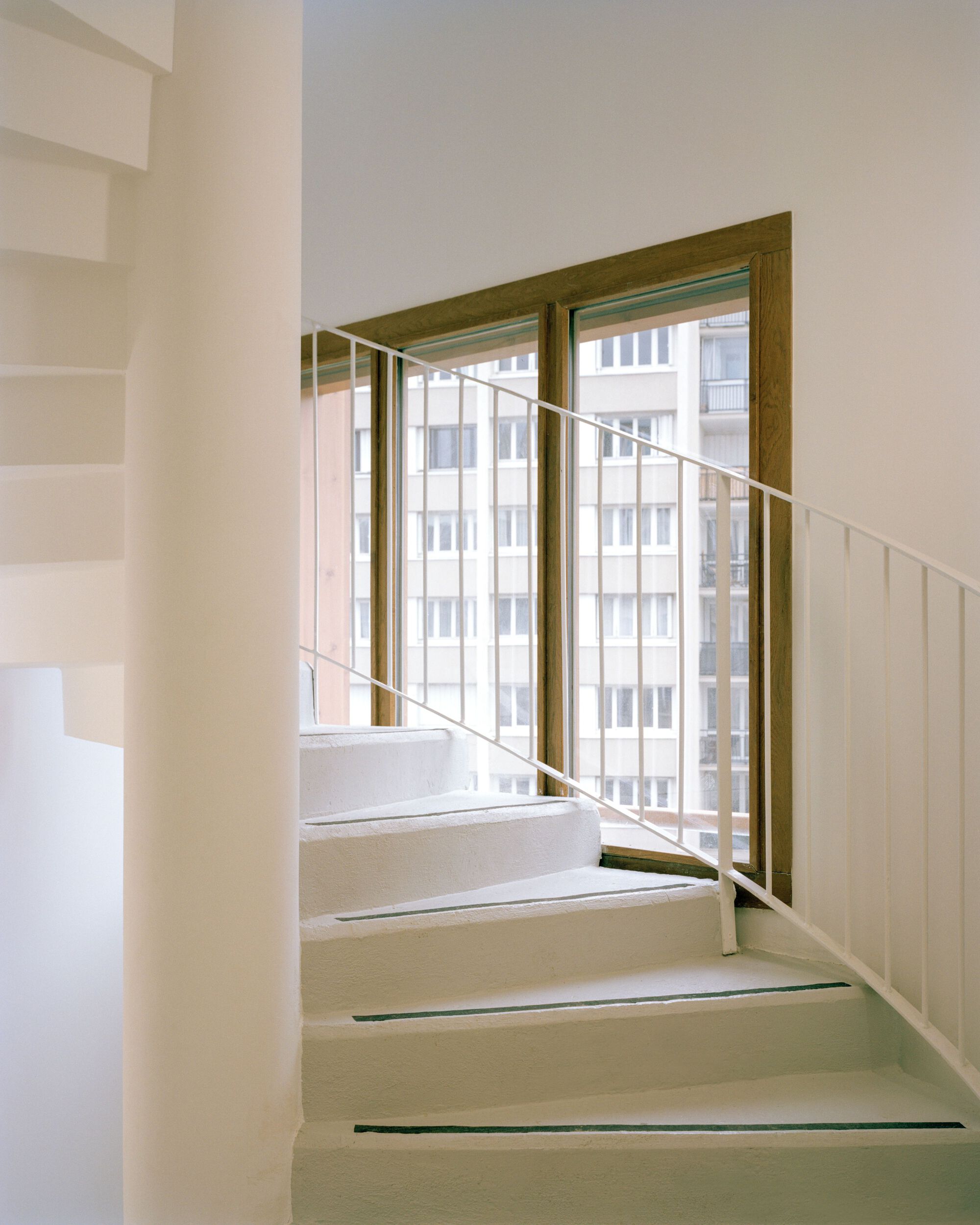 White spiral stair with a thin metal railing rising toward a timber-framed window that looks onto an apartment block
