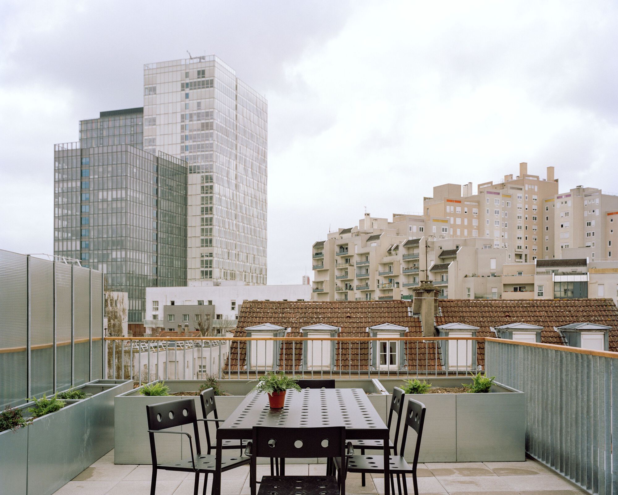 Rooftop terrace with a black metal table and chairs, zinc planters and glass towers rising behind under heavy grey cloud