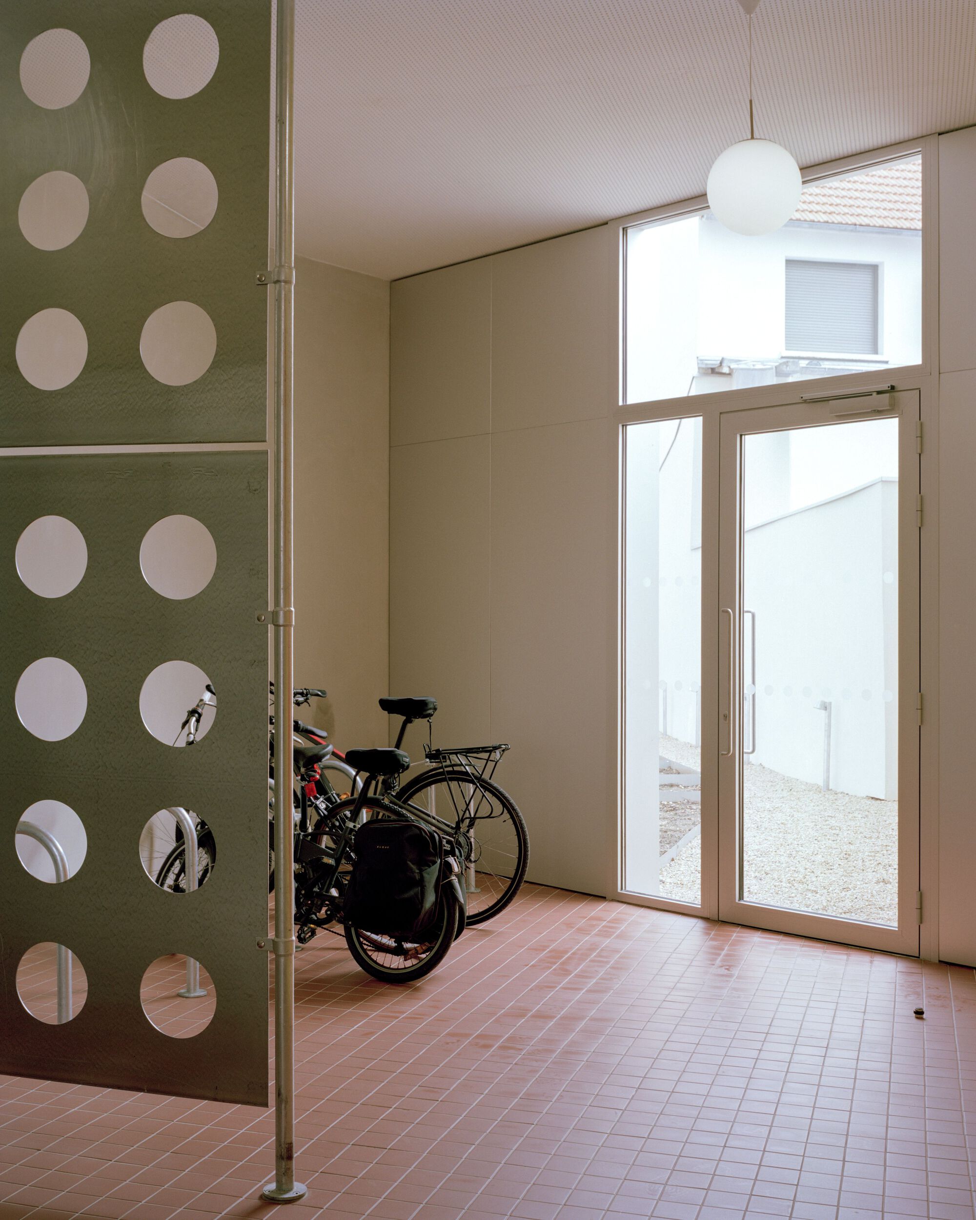 Ground-floor bike room with a perforated green screen, pink tile floor and a glazed door opening to a gravel yard