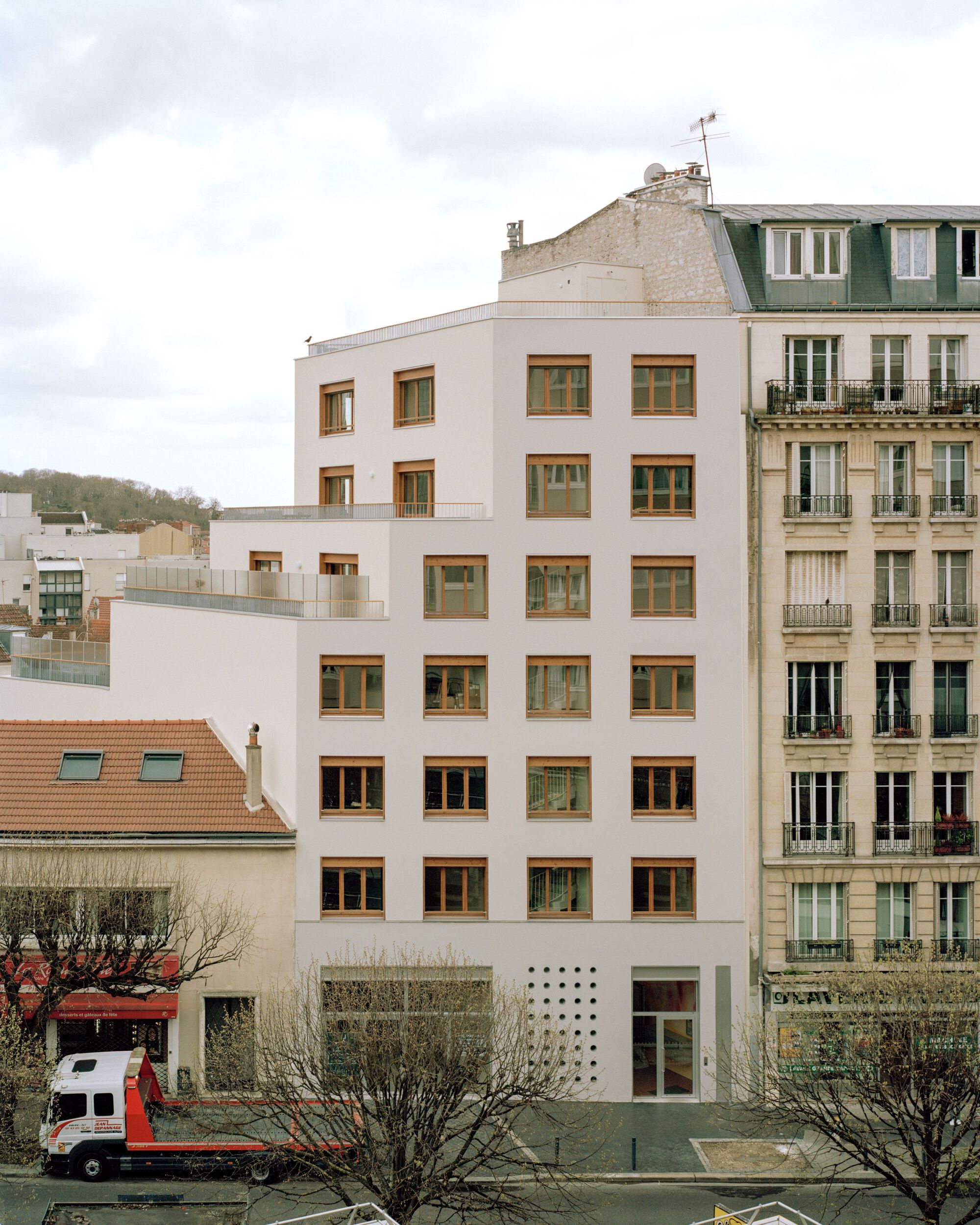 Front elevation of a white block with a grid of timber windows, seen over bare trees and a parked red truck