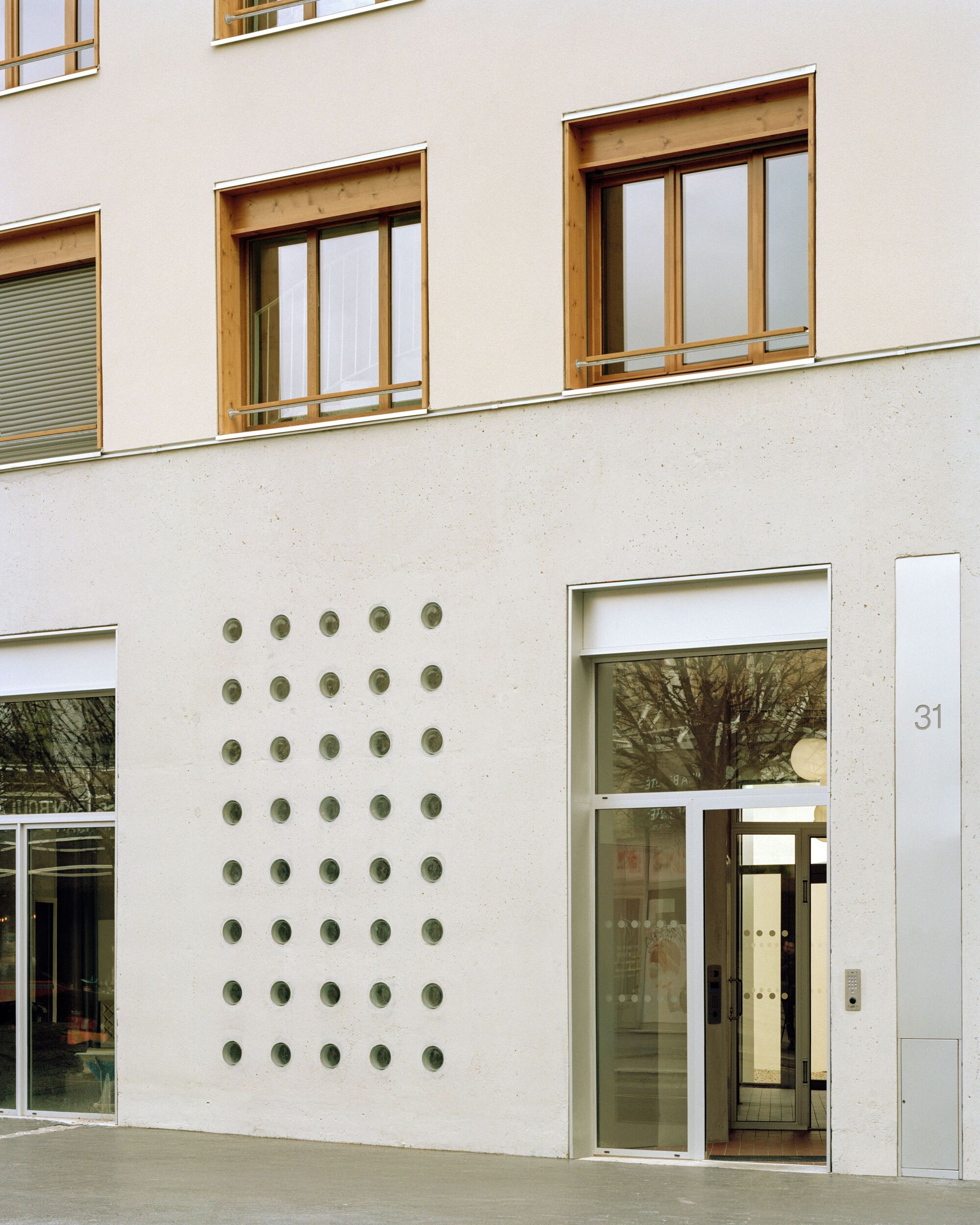 Ground floor facade with a grid of round openings in pale concrete beside a glazed entrance door and timber-framed windows above