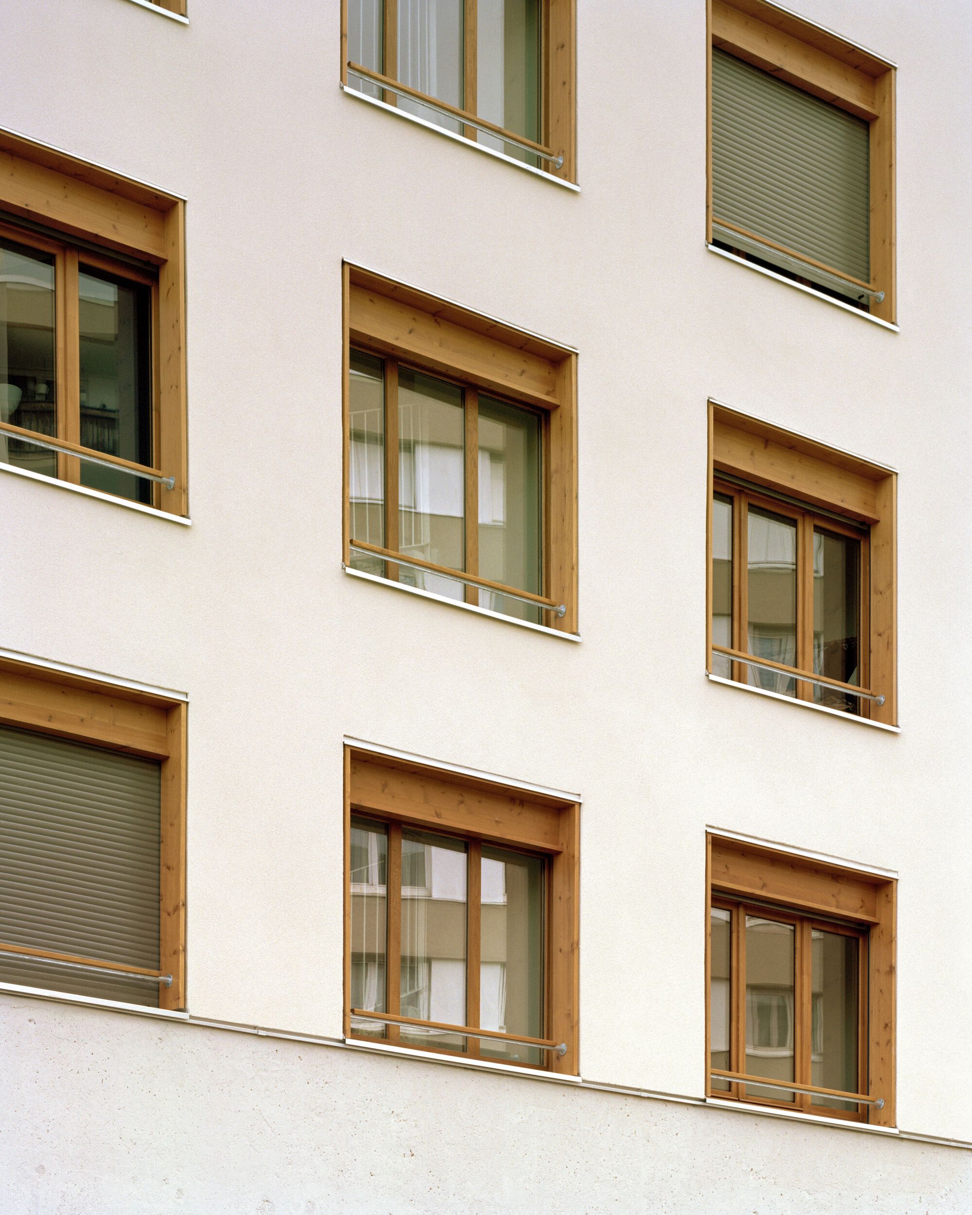 Close view of timber-framed windows with roller shutters set into a white rendered facade under soft overcast light