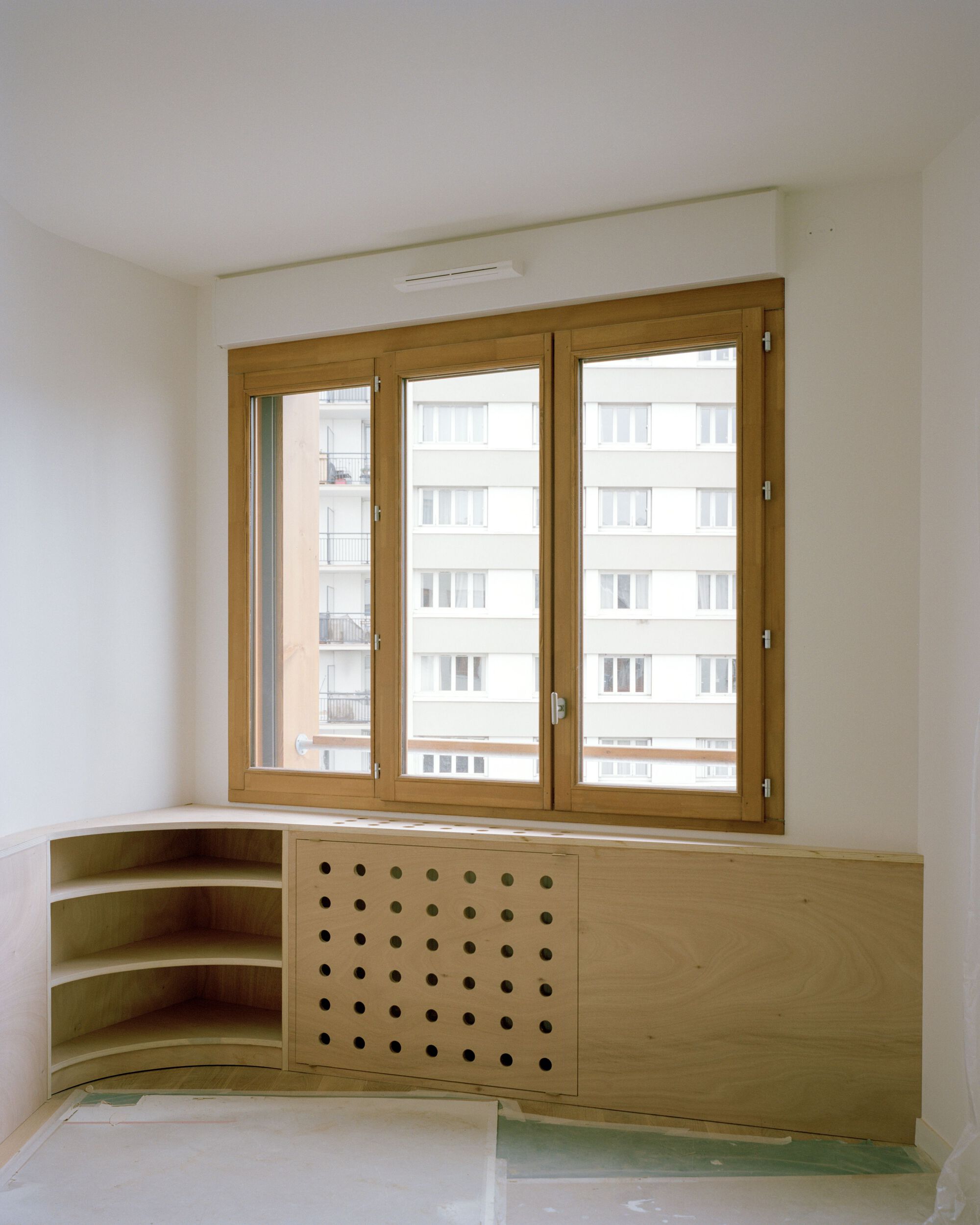 Unfinished room with a timber-framed triple window, curved corner shelves and a perforated plywood radiator cover facing a housing block