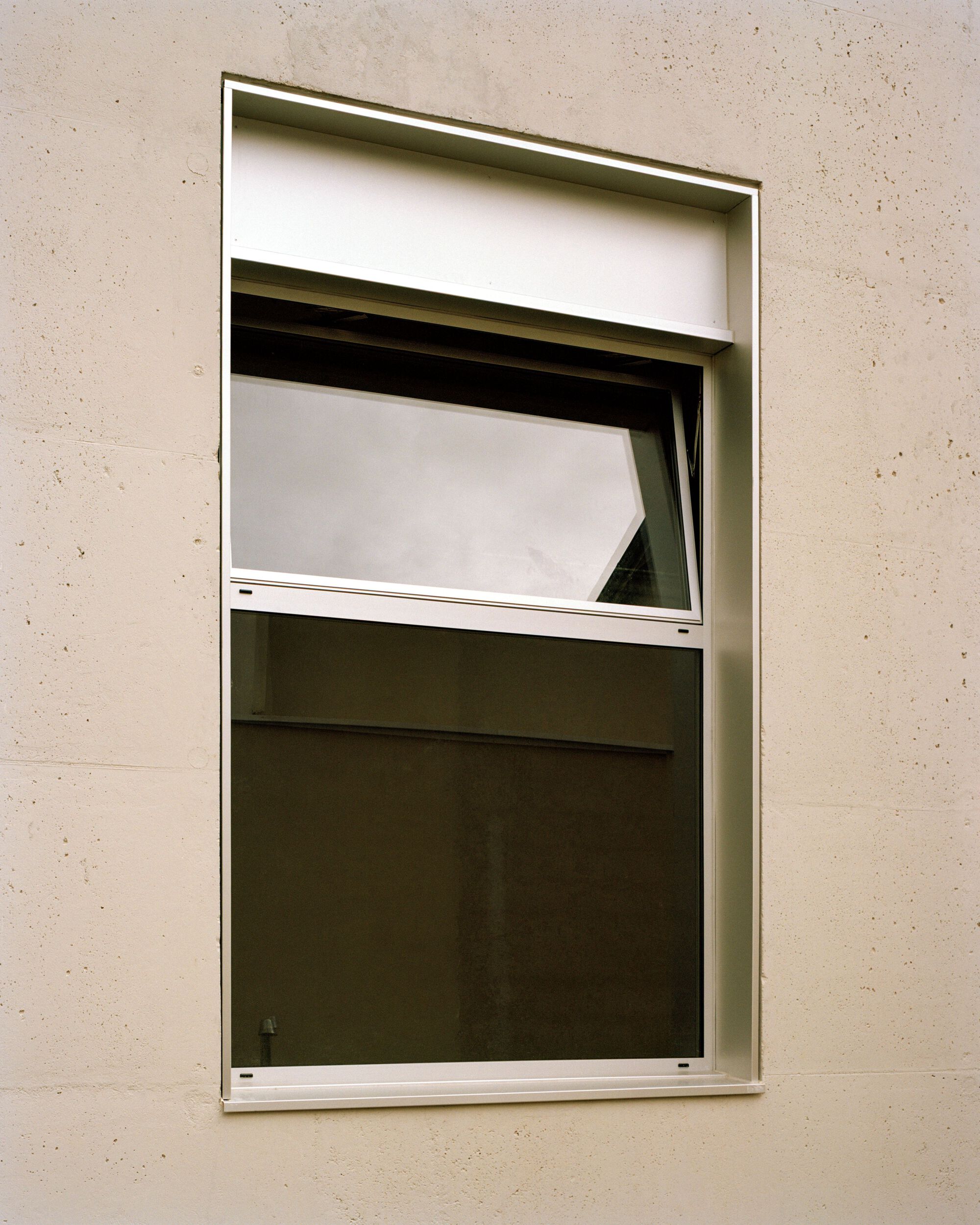 Close-up of a white metal window with a tilted upper sash set into a rough beige concrete wall under overcast light